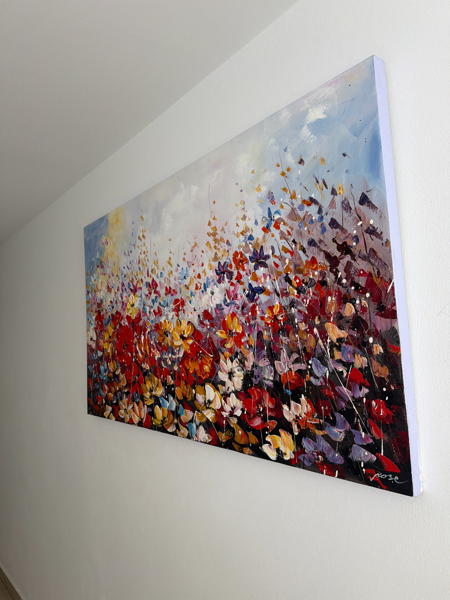 A rectangular canvas artwork featuring a vibrant, abstract floral painting is mounted on a textured white wall. The painting, predominantly displaying shades of red, orange, yellow, blue, and purple, depicts a dense field of flowers with thick, textured brushstrokes. The background of the painting transitions from a bright, hazy sky with hints of yellow and blue to a darker, more muted tone towards the bottom. The artwork is hung slightly askew, with the right side appearing higher than the left. The wall itself is a plain white, with a subtle texture. The lighting appears to be natural, coming from an unseen source, casting a soft illumination on the scene. The overall environment suggests an indoor setting, possibly a home or gallery, in the city of Ayia Napa, Cyprus. There are no people visible in the image.