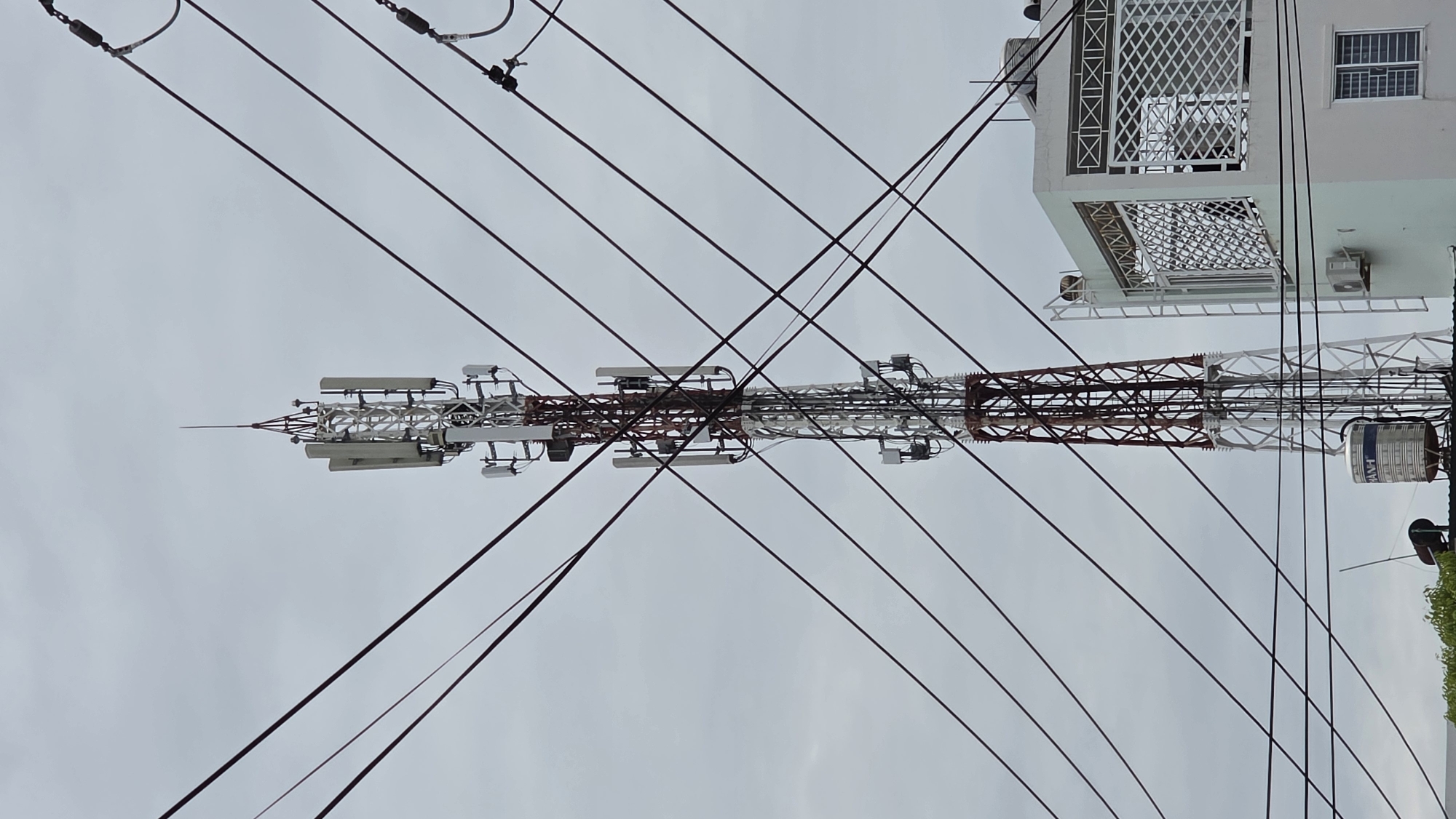 The image captures a dense network of urban infrastructure, dominated by a tall communication tower under an overcast sky. The tower, a metal lattice structure, is painted in sections of white and reddish-brown. Numerous platforms extend from the tower, laden with various communication equipment, including multiple rectangular panel antennas, cylindrical components, and a complex web of cables. A sharp, pointed lightning rod sits atop the tower.
In the foreground, a dense array of dark power lines or communication cables crisscrosses the frame, partially obscuring the tower and creating an intricate, overlapping pattern.
To the right of the tower, part of a white multi-story building is visible. It features a balcony with a distinct white metal lattice railing and security grilles on its windows. An outdoor air conditioning unit is mounted on the side of the building. A blue cylindrical water tank, bearing the text "HAVI," is positioned near the base of the visible building section. Green foliage is partially seen at the bottom right.
The sky is uniformly overcast, indicating a cloudy daytime setting. The scene depicts a functional, built environment with significant technical infrastructure. The image is presented rotated 90 degrees clockwise from its original orientation.