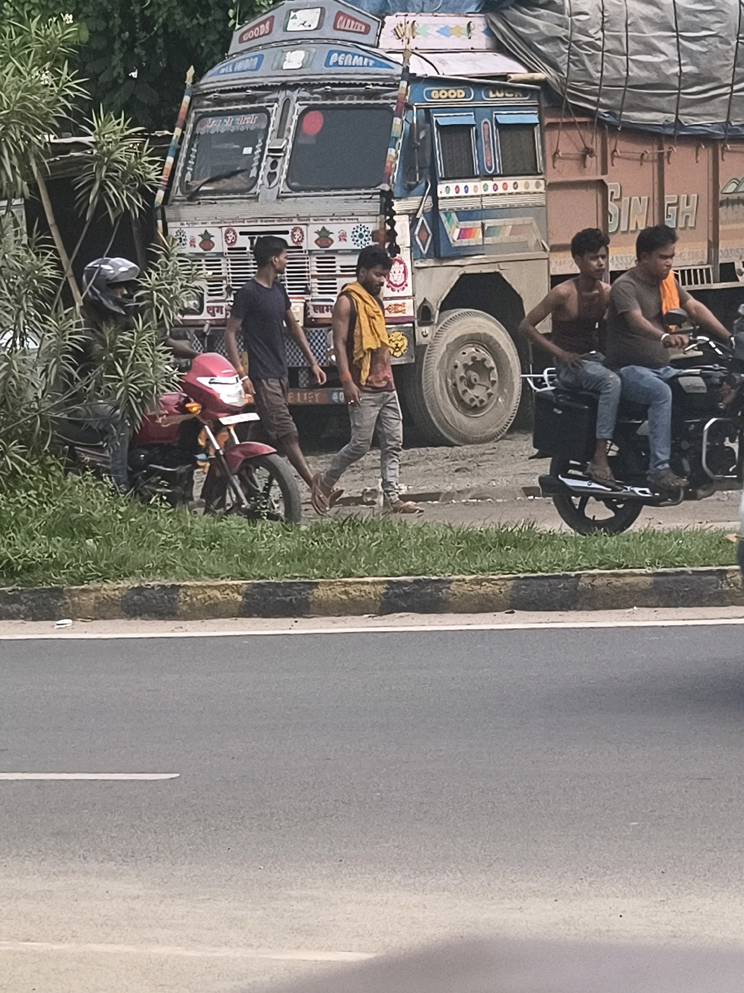 The image depicts a street scene in Purnia, India, characterized by a large truck and several individuals. The truck, a prominent element, is a heavy-duty vehicle with colorful branding and a covered cargo area. In front of the truck, three men are visible. One man, wearing a dark t-shirt and shorts, stands to the left. Next to him, another man in a tank top and jeans is seated on a motorcycle. A third man, wearing a light-colored t-shirt and jeans, is riding on the back of the same motorcycle, holding an orange cloth. Another man, dressed in a yellow garment, is walking away from the motorcycle towards the truck. To the left of the truck, a red motorcycle is parked amongst lush green foliage. The ground appears to be paved, with a distinct yellow and black striped curb separating the road from the grassy area. The overall environment suggests a roadside setting within a city.