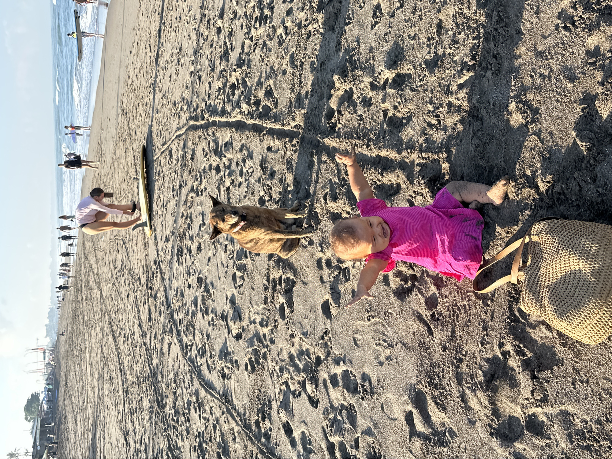 A low-angle, eye-level outdoor shot captures a beach scene in North Kuta, Indonesia. In the foreground, a baby sits in the sand wearing a bright pink dress and appears to be playing. To the baby's left is a woven straw bag. Behind the baby, a medium-sized brindle-colored dog sits attentively, looking forward. In the middle ground, a person in a light pink long-sleeved top and dark bottoms is bent over a yellow surfboard on the sand. Further in the background, numerous people are walking along the beach, some near the water's edge. The ocean with gentle waves is visible in the distance, and the sky is clear and bright. The sandy beach is marked with footprints and tire tracks.