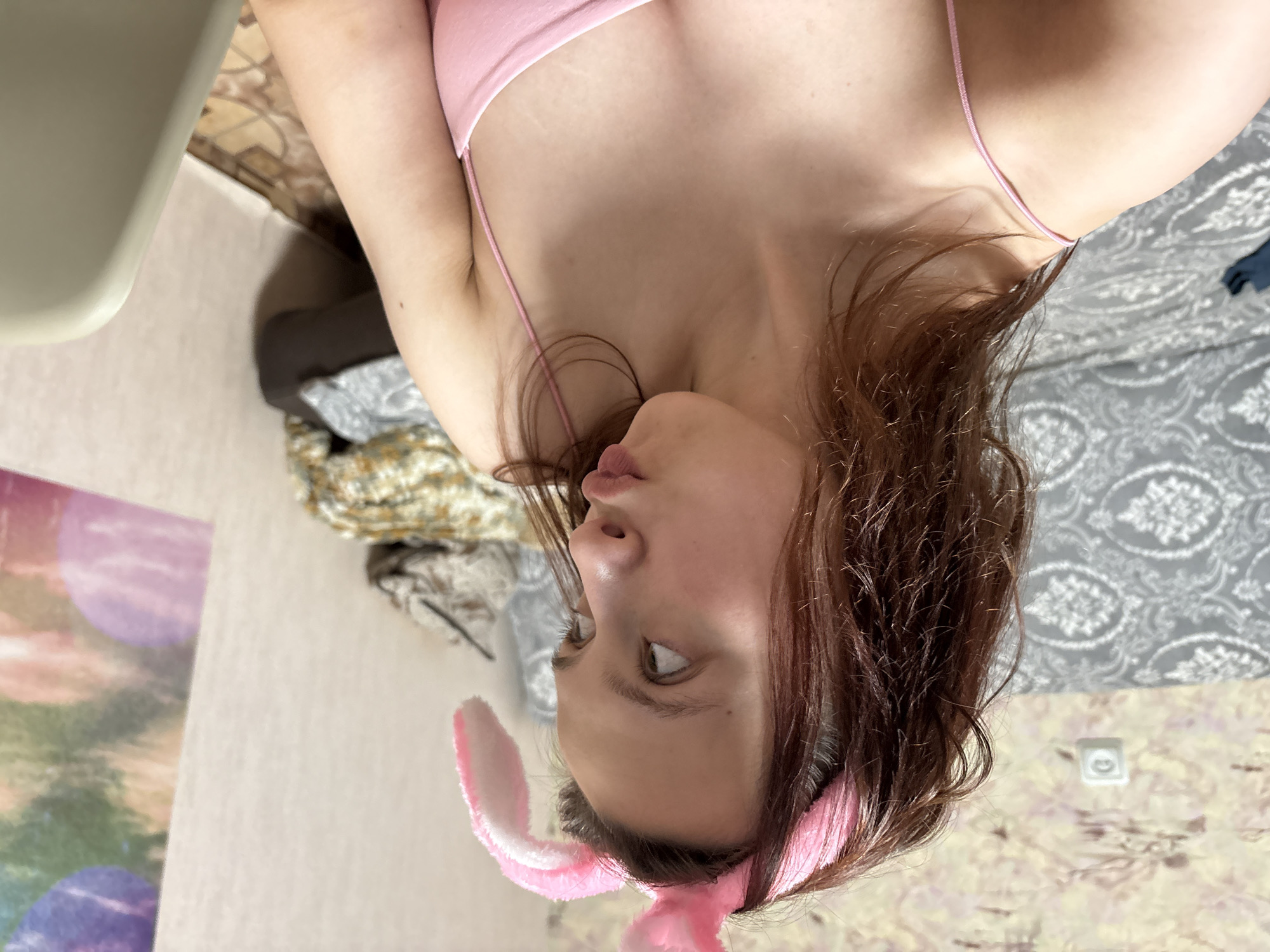 A young woman with shoulder-length reddish-brown hair is the central focus of this indoor portrait. She is wearing a light pink spaghetti strap top and a pink bunny ears headband. Her head is tilted slightly, and she looks upwards and to her right with a pensive or thoughtful expression. Her lips are slightly parted, and her eyes appear to be a light color.
The setting is an indoor room, likely a bedroom. Behind her, a grey duvet or blanket with a white floral or damask-like pattern is visible, suggesting a bed. To the left, there's a glimpse of a light-colored object, possibly a chair or table, and a wall with a subtle textured pattern. Further left, a colorful, abstract poster or picture featuring purple and green tones hangs on the wall. In the background, a white electrical outlet is mounted on a patterned wall. Some other fabrics or clothing items are casually piled up in the mid-ground. The lighting suggests it is daytime. The overall scene conveys a relaxed, candid moment within a domestic environment in Прикамский, Russia. There is no visible text in the image.