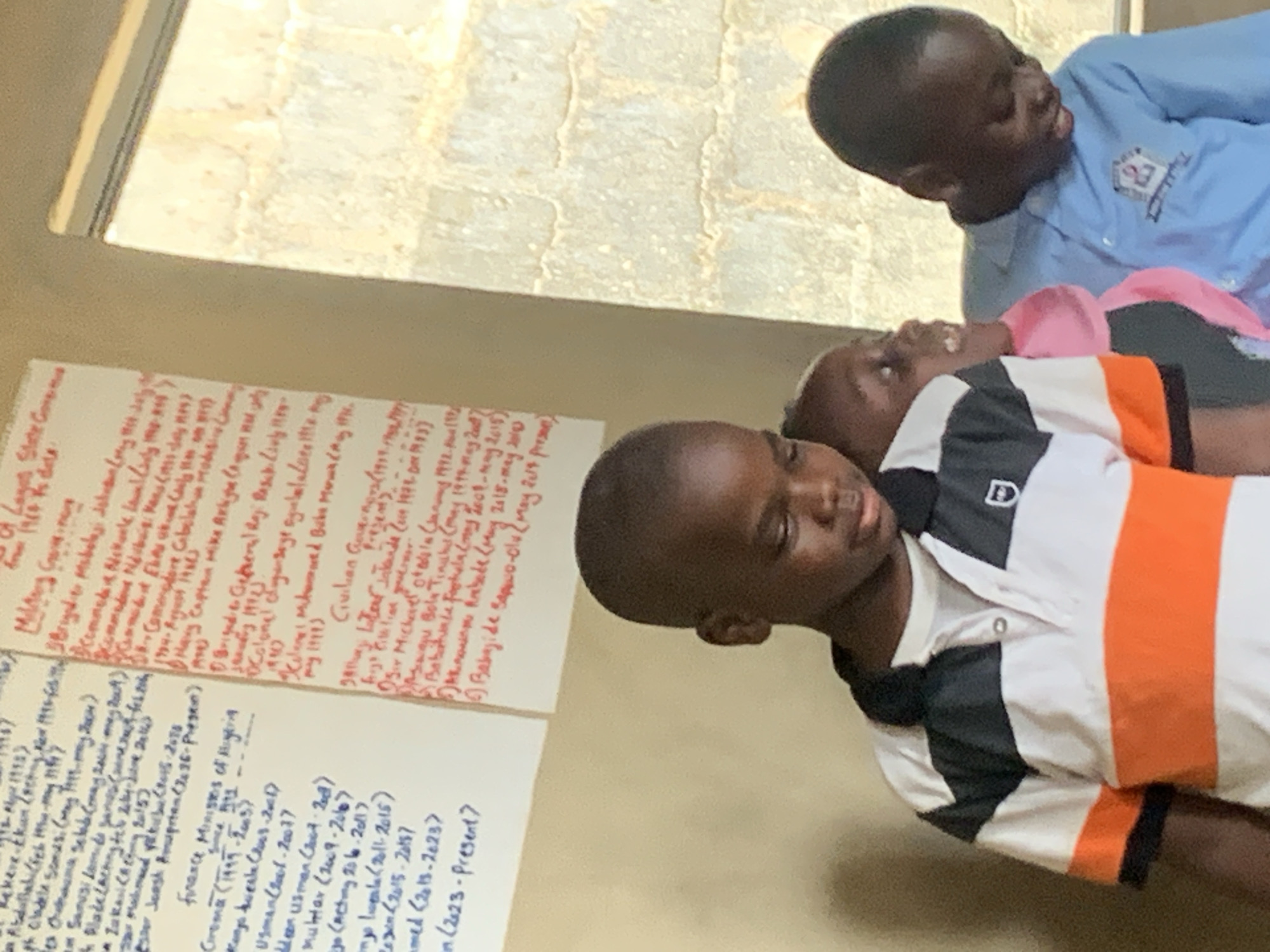 Here is a description of the image:
The image shows three Black children indoors. They are standing next to a wall on which two white posters are hung. The posters are covered in text, and some of the text is highlighted in red ink. The text appears to be a list of governors or other political figures and the dates they served. One child is wearing a white polo shirt with a black collar and a thick orange stripe across the chest. He has a "D" logo on the left side of the collar. Another child is wearing a light blue shirt. The third child is wearing a pink or red top and appears to be lying down with the other two standing beside her.