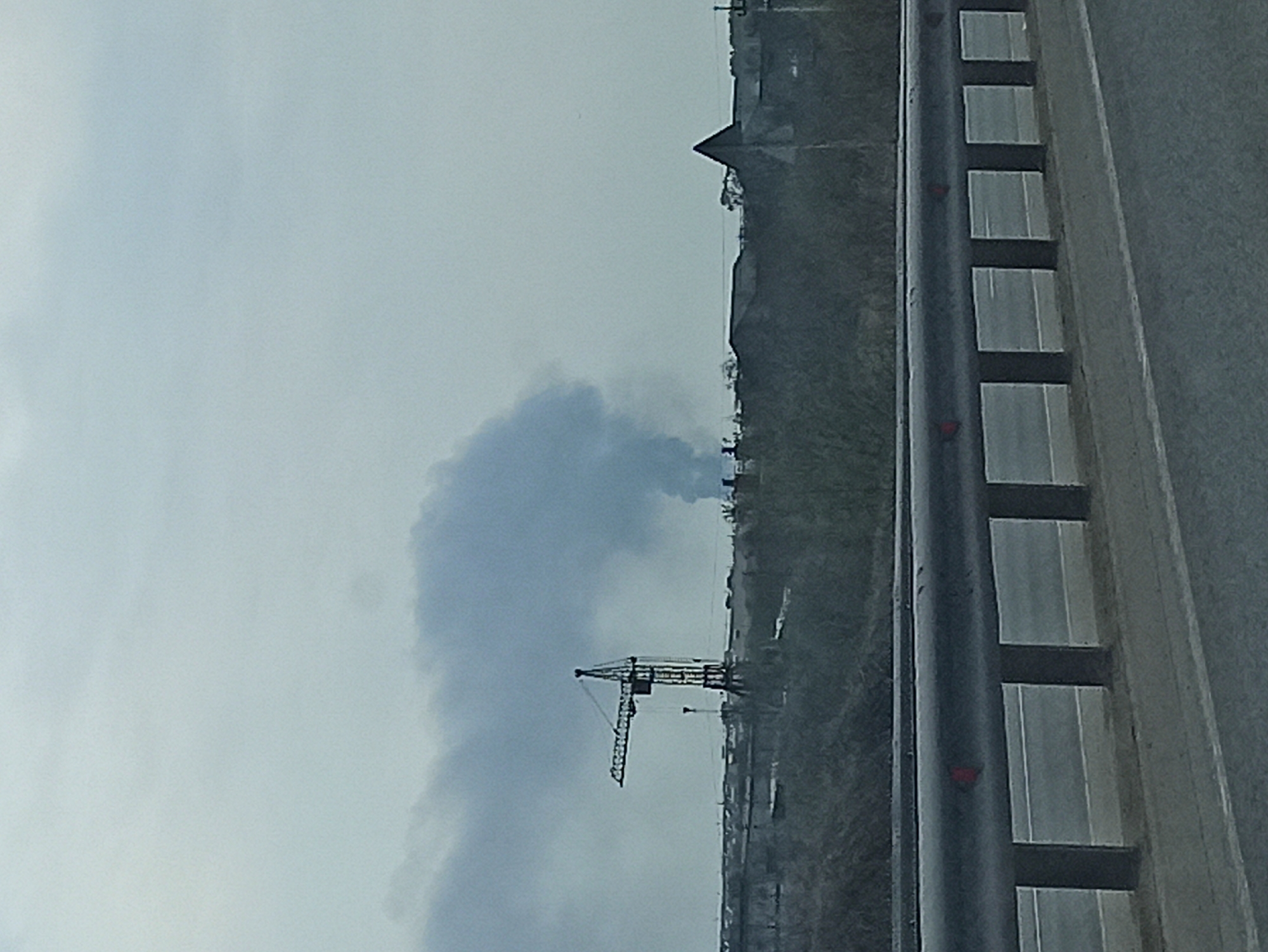 The image shows a view from a bridge or elevated structure in Kitsjanovo, Russia. Visible in the foreground is a section of the bridge's railing. In the middle distance, a construction crane is silhouetted against a cloudy sky. Plumes of smoke are visible behind the crane, possibly indicating an industrial area or event. A landscape with a church or tall building on a small hill is visible further back.