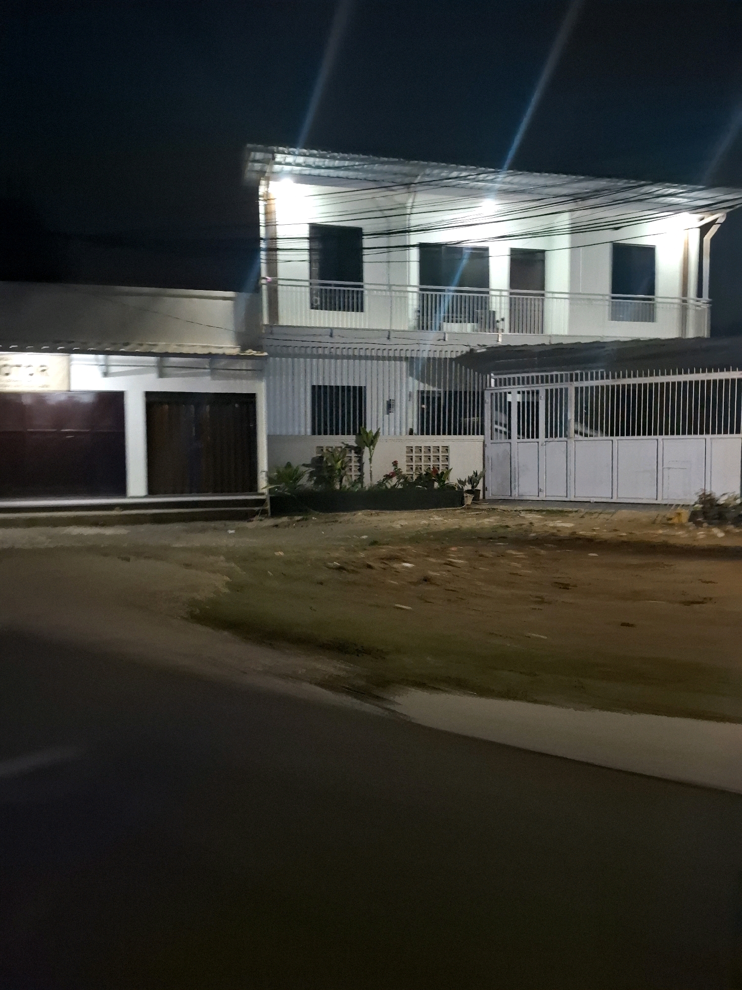 The image depicts a two-story building at night, illuminated by artificial lights. The building appears to be made of concrete and has a corrugated metal roof. The lower level features dark-colored double doors, possibly for a garage or storefront. The upper level has a balcony with a white railing and several windows. Electrical wires are visible strung across the building and the sky. To the right of the building, a white metal gate provides access to a fenced area. The ground in front of the building is uneven and appears to be dirt or gravel, with some sparse vegetation and plants. A paved road is visible in the foreground, suggesting the building is situated along a street. The scene is devoid of any people. The location is identified as Ciheulang Tonggoh, Indonesia.