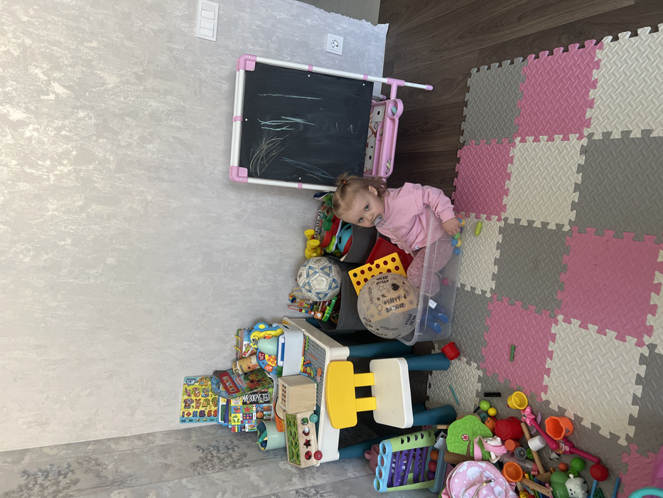 A young child's play area is depicted in an indoor setting within the city of Leninakan, Russia. A toddler, with light hair and a pacifier, wearing a pink long-sleeved top and patterned pants, is seated inside a clear plastic storage bin. The child looks directly forward with a neutral expression. The room features light grey textured wallpaper on the walls and a floor composed of brown wood laminate transitioning into interlocking foam mats in pink, grey, and off-white.
To the left, a child-sized blue and white table with a yellow chair is positioned against the wall, topped with a wooden sorting toy, various books, and smaller toys in white and green containers. A collection of diverse toys, including building blocks, plastic figures, and a musical toy, is scattered across the foam mats in the foreground. Behind the child, two grey fabric bins hold additional toys, including a white and black soccer ball. The clear plastic bin containing the child also holds a large transparent balloon with text and a yellow plastic toy. To the right, a pink and white child's easel with a chalkboard surface covered in white scribbles stands against the wall, adjacent to a white electrical outlet and a light switch plate.