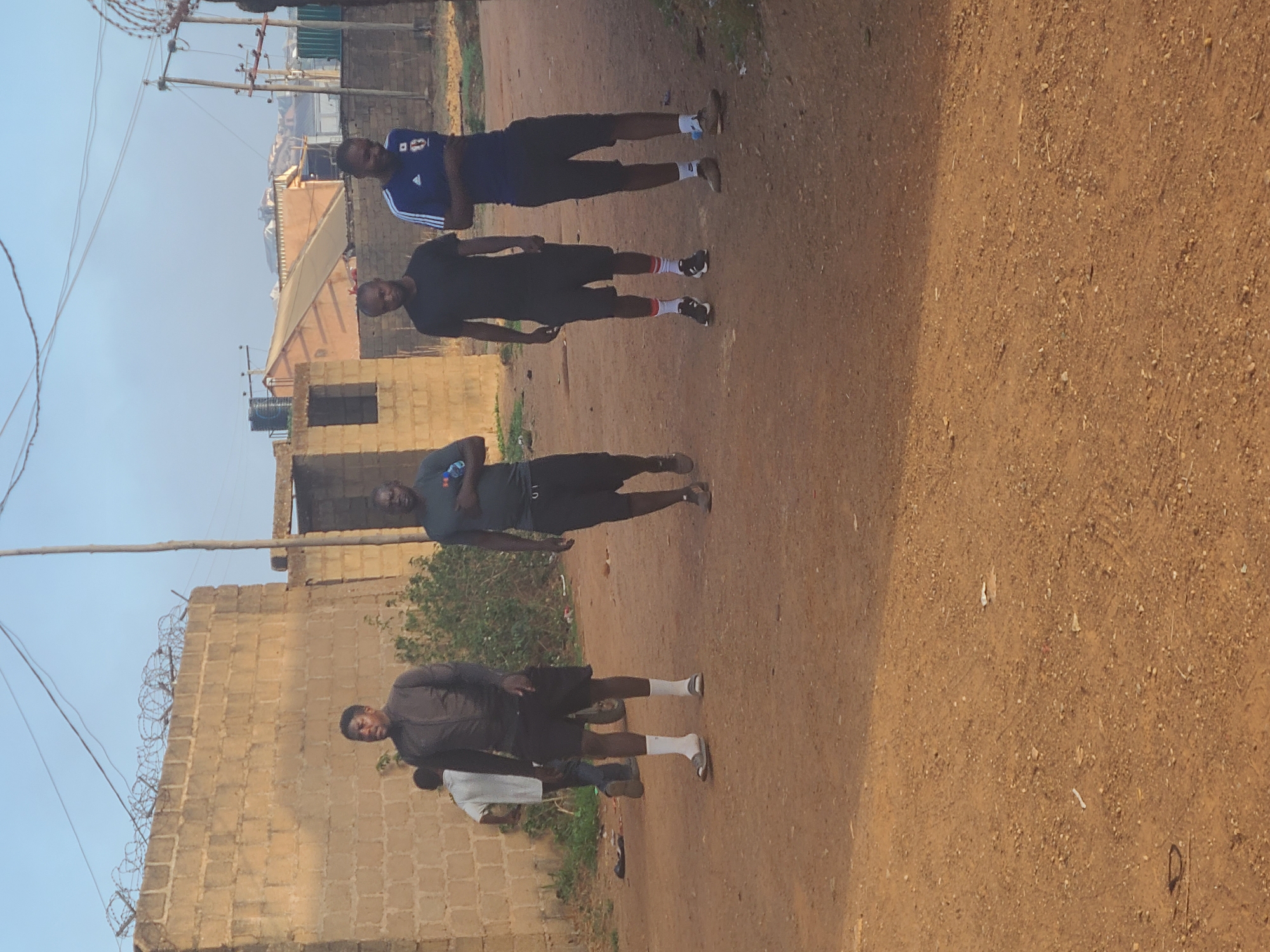The image depicts four men standing on a dirt road. The man furthest from the camera is wearing a blue and white sports jersey with dark shorts. The next man is wearing all black. The third man is wearing a grey t-shirt and dark shorts. The closest man to the camera is wearing a grey jacket and dark shorts with white socks. All four men appear to be of African descent. The background includes buildings made of stone and a blue sky. Based on the appearance of the buildings and the men, this scene likely takes place in Jos, Nigeria.