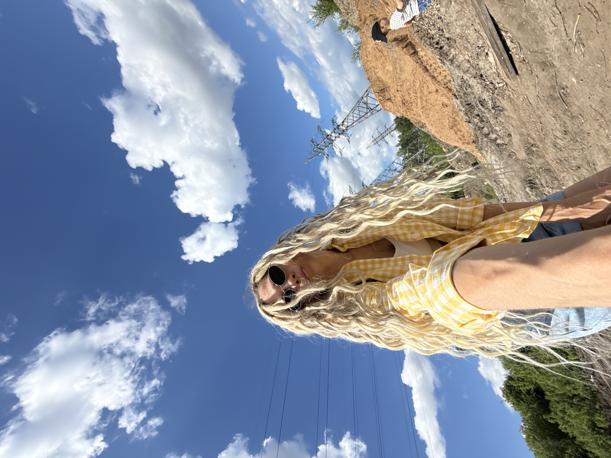 A woman with long, wavy blonde hair is photographed from a low angle against a bright blue sky with scattered white clouds. She wears round sunglasses and a yellow-and-white checkered shirt over a white top, with denim shorts visible. Power lines and electrical towers are in the background, along with a dirt embankment. There is another person sitting on the embankment. The location is near the city of Заволжье, Russia based on the power lines.
