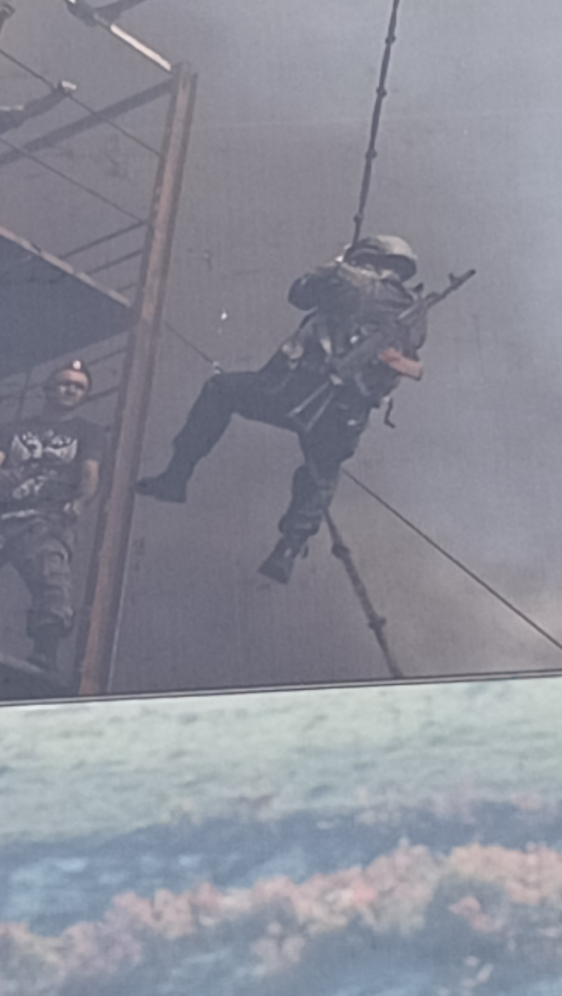The image depicts a dynamic scene of a military or tactical demonstration, likely taking place outdoors against a smoky or hazy backdrop. The central figure is a person in full tactical gear, actively rappelling down a thick rope from an elevated structure. This individual is clad in a dark uniform, helmet, and tactical vest, and holds an AK-pattern assault rifle, possibly an AK-74, in a ready stance.
To the left, a second man stands on a wooden or metal scaffolding, observing the rappelling activity. He wears a black T-shirt featuring a white graphic (possibly a wolf or dog head), camouflage trousers, and a headlamp on his forehead. He has a slight smile, suggesting a relaxed atmosphere for the event, perhaps indicating an instructor or demonstrator.
The lower portion of the image shows a blurry, abstract landscape in hues of green, blue, and reddish-brown, which appears to be a painted backdrop or a simulated environment, further suggesting an exhibition rather than a real-world operational setting. The overall ambiance created by the haze and the activity points to a public display or training exercise of tactical skills. Located in Tiraspol, Moldova, this scene could represent a demonstration by local security forces or a similar organization.