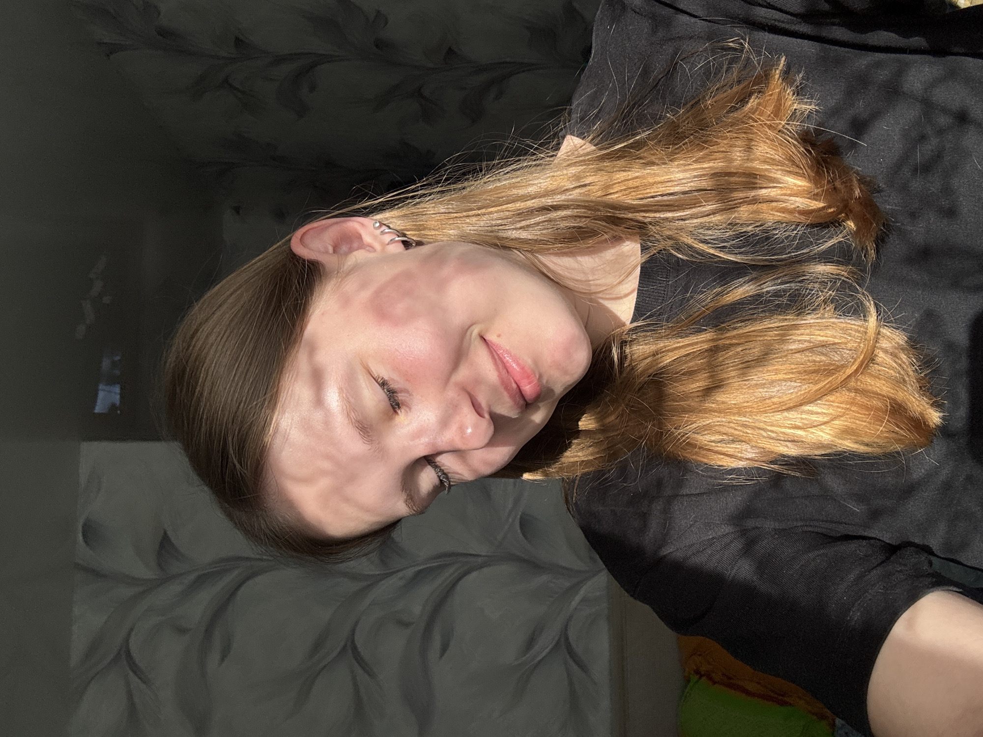 A young woman with auburn hair, wearing a black top, lies on a grey, patterned surface. The woman is smiling slightly with her eyes closed. The scene is illuminated with dappled sunlight. No city location (Krilovo, Russia) is discernible from the image.