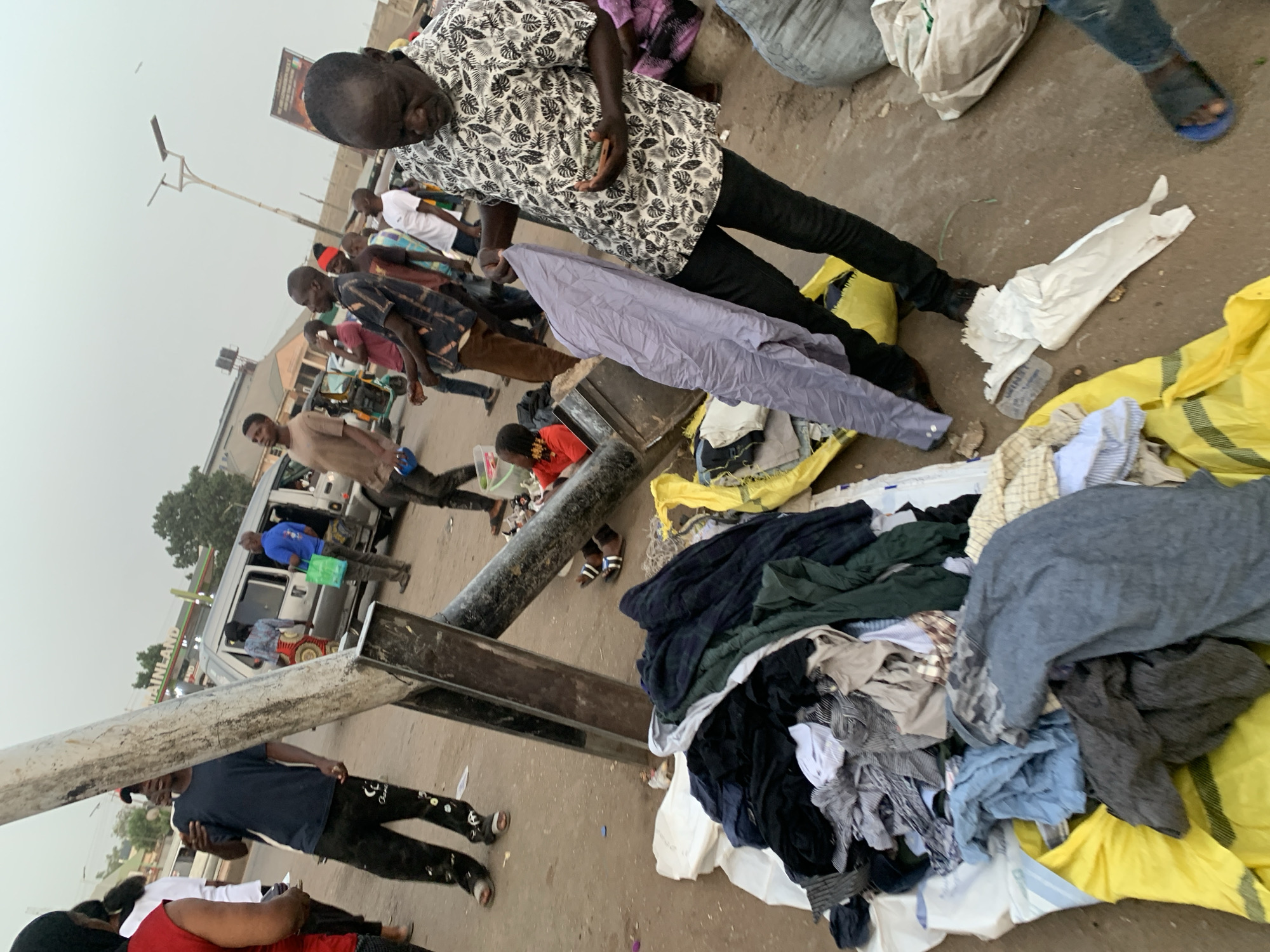 The image captures a bustling street scene, likely a market, under an overcast sky in Jos, Nigeria. The photograph is taken from a slightly elevated perspective, looking down and across a street where numerous people are gathered.
In the immediate foreground, a man wearing a black and white patterned shirt and black trousers stands, looking down at large piles of assorted clothing spread out on yellow and white tarps or bags on the dusty ground. A light purple garment is draped over his left arm, suggesting he is either sorting or displaying the clothes for sale. His hands appear to be holding something small, possibly a phone or a small item from the merchandise.
To the right of this man, more piles of clothes are visible, featuring a mix of shirts, trousers, and other fabrics in various colors like grey, blue, black, and green. The ground around the clothing is littered with some debris, including what looks like a crumpled white bag and a plastic bottle.
Further into the midground, several other individuals, mostly men, are present. Some are walking, while others appear to be standing or interacting. One man in a black t-shirt and black pants with white patterns on the legs is partially visible, and a child in a red shirt is also seen crouching near the ground. A long, dark metal bar or pipe lies across the scene, creating a visual barrier.
In the background, a white van or bus is parked, with people near it, and parts of buildings are visible. A street light pole stretches upwards, and a large sign with some unreadable text is partially seen above the distant buildings. Another green sign, possibly for a business, has the word "MAINLAND" visible on it. The overall impression is one of an active urban market environment during the daytime, characterized by informal vending and a lively crowd.