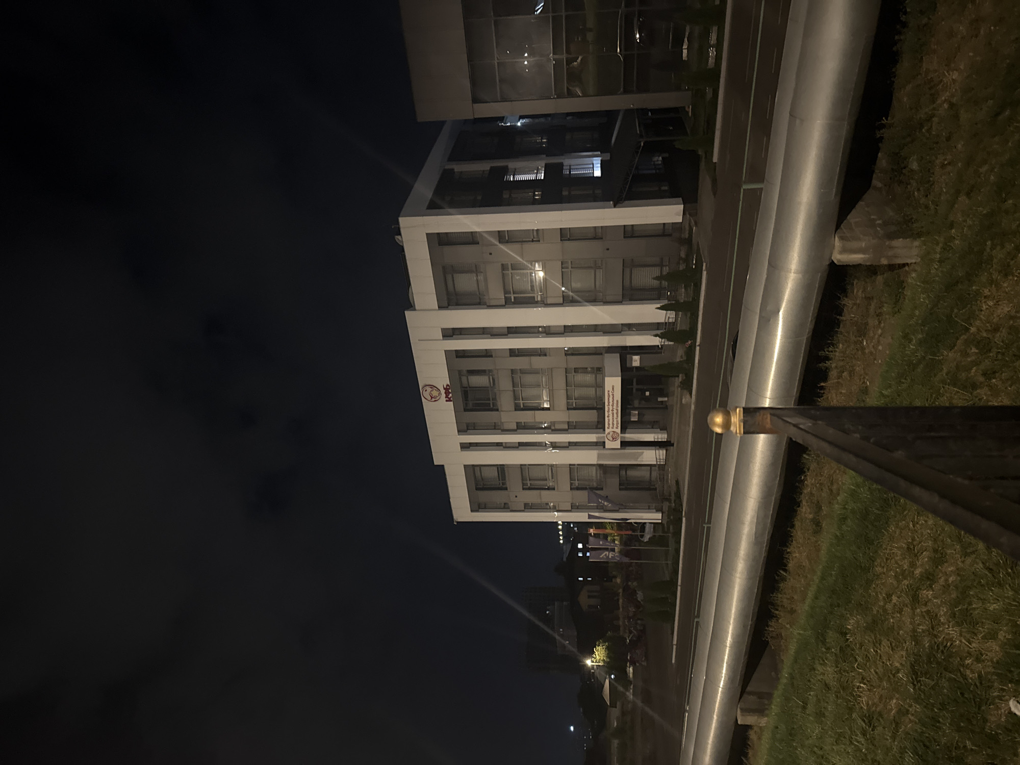 The image shows the building of the Kyrgyz Football Union in Bishkek, Kyrgyzstan, at night. The modern, multi-story building is illuminated, with the logo of the union visible on the facade. The surrounding area appears to be a commercial or administrative district, with a road and large pipes in the foreground. The sky is dark, indicating nighttime, and there are some clouds visible. The overall scene suggests an urban environment at night.