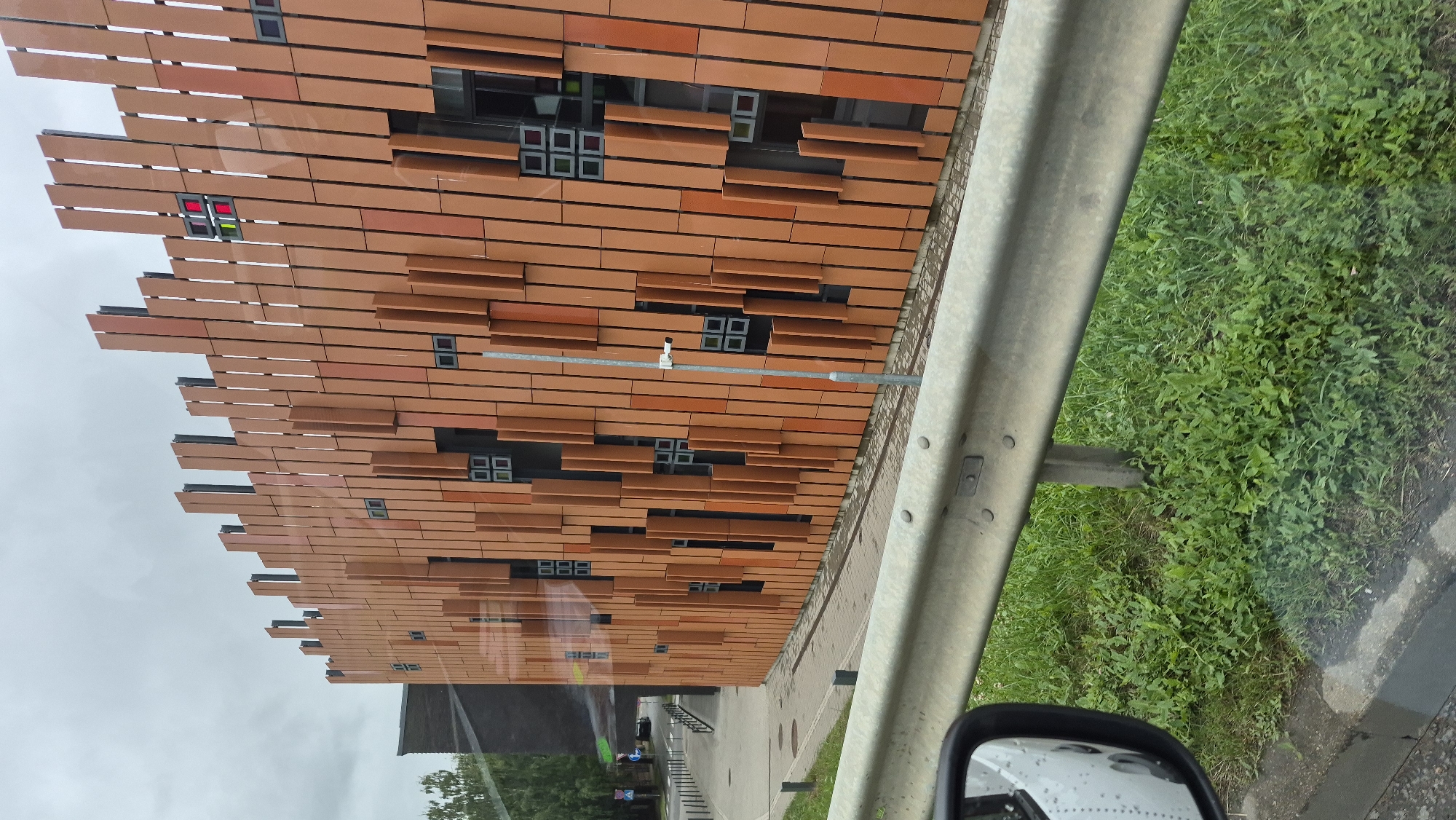 The image shows the facade of a modern building with a distinctive design. The building features a curved wall clad in vertical, terracotta-colored panels of varying lengths and depths, creating a textured and dynamic surface. Small, dark, square window openings are visible, some of which contain colored glass, including red and green panes. A silver-colored security camera is mounted on a pole in front of the building. To the left, a portion of a dark, brick-clad building is visible, along with trees and a street with a sidewalk. In the foreground, a metal guardrail runs horizontally across the frame, with green grass and vegetation visible below it. The scene appears to be captured from a moving vehicle, as indicated by the reflection in a car's side mirror on the left side of the image. The sky is overcast. This is the central entrance of the Daugavpils Olympic Centre in Daugavpils, Latvia.