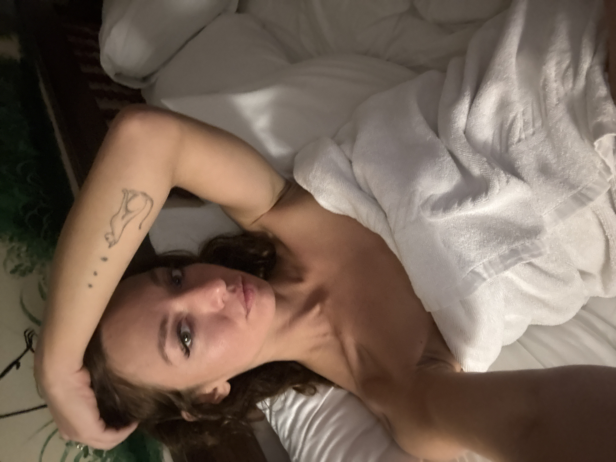 A fair-skinned individual with brown hair is lying in a white bed, partially covered by a white towel or blanket. The person is facing the camera with their left arm raised, displaying a tattoo of a cat and three small dots on their forearm. The background shows part of a dark wood headboard and a green patterned surface, likely a wall covering or decor. The setting is indoors, with soft lighting suggesting it is either early morning or evening. There is no information indicating the location is in Đà Nẵng, Vietnam.