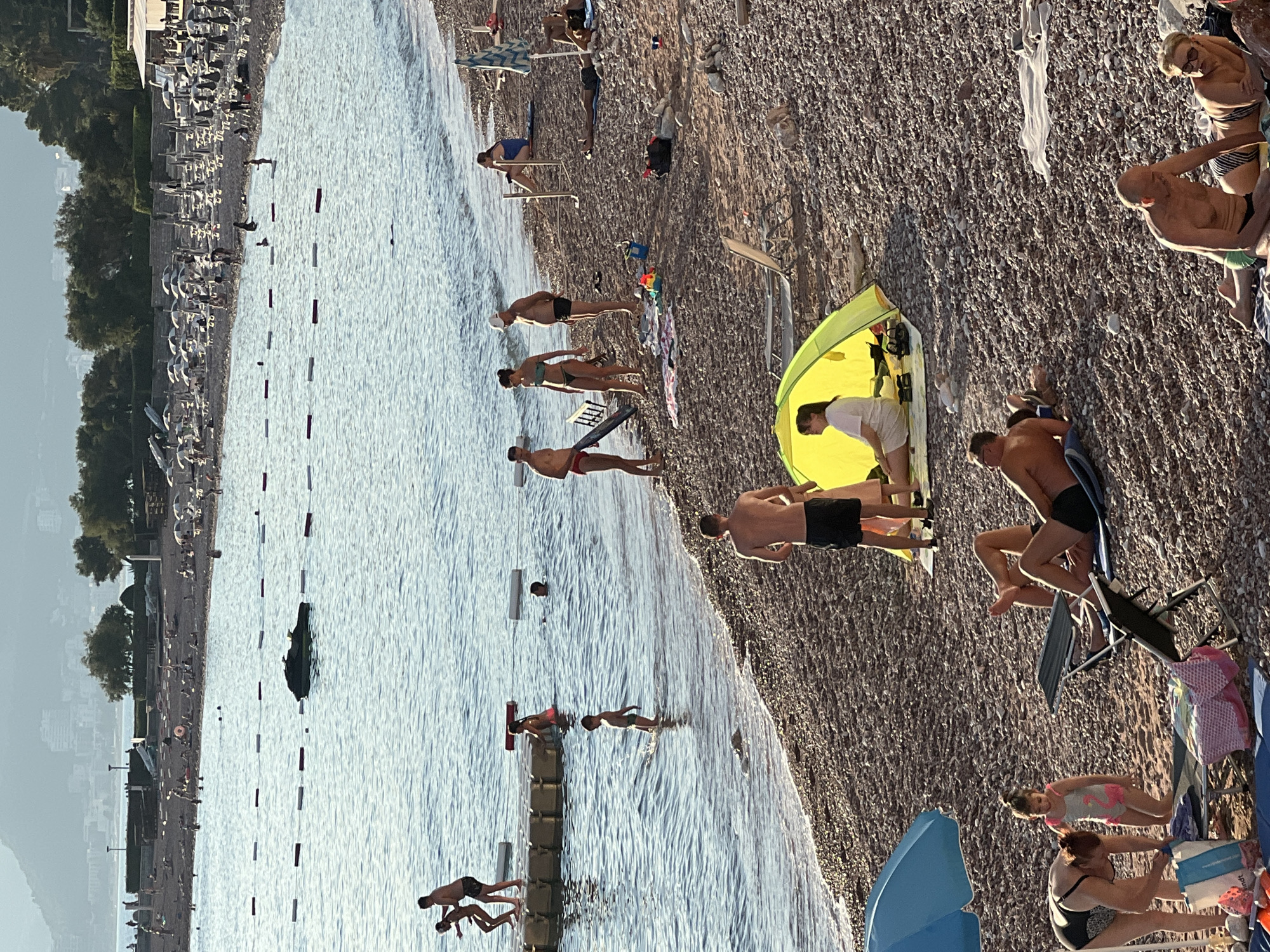 In Sveti Stefan, Montenegro, a crowded pebble beach is depicted with many people enjoying the sun and sea. In the foreground, a woman in a black and white swimsuit is bent over, placing items into a bag while a young girl in a flamingo-patterned swimsuit stands beside her. Several beach chairs are set up on the shore. Further back, a yellow tent is set up on the beach with a woman sitting inside. Men and women are scattered across the beach, some sunbathing on towels, others standing and talking.
In the water, several individuals are swimming and playing. Two children are standing on a floating platform in the sea, with another child wading nearby. Further out, a jet ski is visible on the water. The sea is calm, and a line of buoys marks a swimming area. In the background, rows of lounge chairs and umbrellas are set up on the beach, and a town with buildings and trees is visible on the hillside. The overall scene is one of a bustling summer day at a popular tourist destination.
