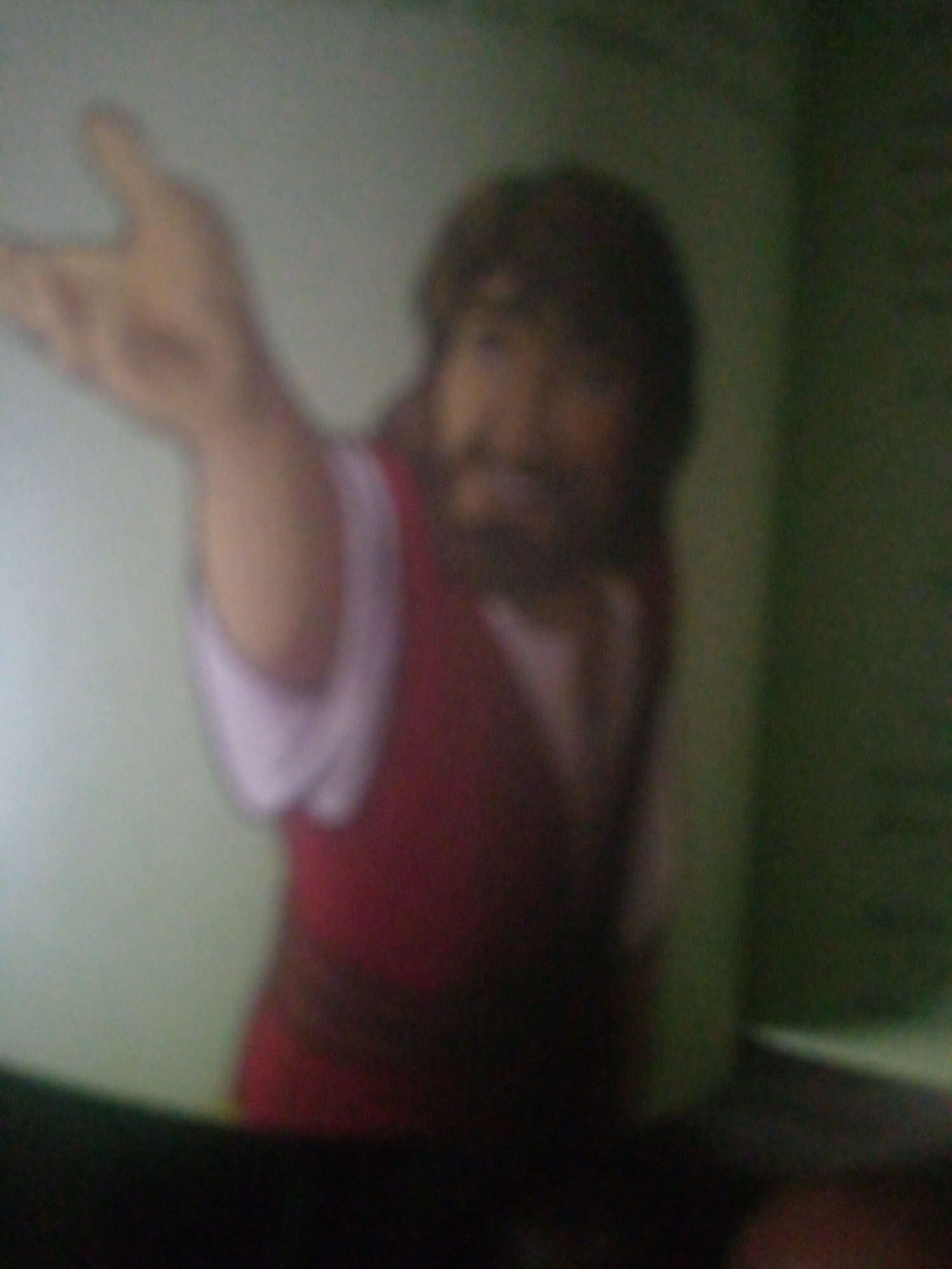 The image, taken in Jauro Yinu, Nigeria, depicts a blurred figure of a person with a beard, wearing a red garment over a white undershirt. The person's left arm is raised, with their hand open and extended outwards. The background is a mix of light green and dark green, with some darker areas at the bottom suggesting a surface. The lighting is dim, contributing to the overall blurriness of the scene.