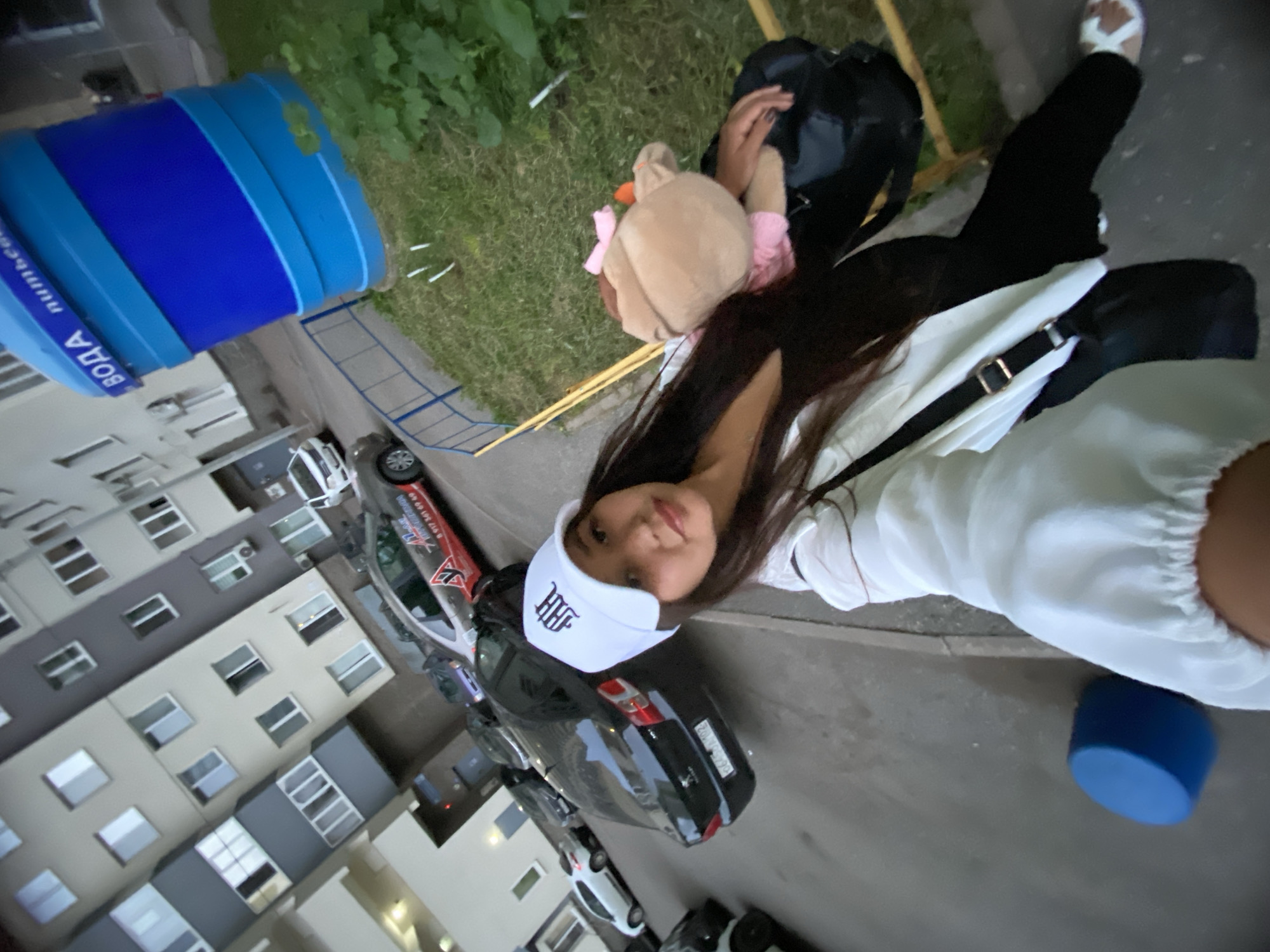 A young woman in a white baseball cap and white jacket stands on a paved surface in front of a multi-story apartment building. She holds a stuffed toy and a black bag. Cars are parked on the street. To the right of the woman, a large blue water tank stands on a grassy area next to a metal fence. The building has a facade of light and dark gray panels with many windows. The scene is likely in a residential area of Ufa, Russia, given the inscription "ВОДА" (Water) on the blue tank, which is a common sight in Russian cities, and the style of the apartment buildings.