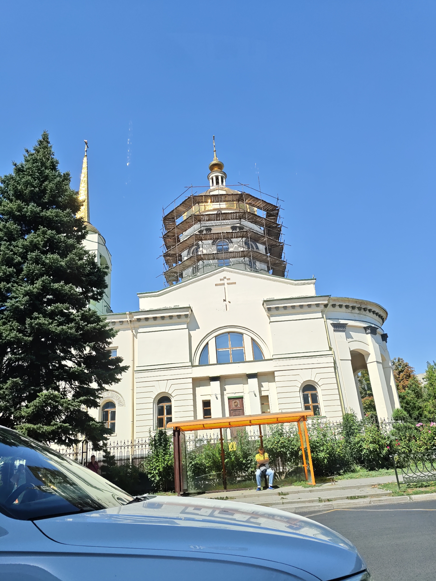 The image depicts a daytime view of the ascent to the Holy Trinity Cathedral in Rostov-on-Don, Russia, as seen from a vehicle. The cathedral's main dome is under scaffolding, indicating ongoing construction or renovation. A tall evergreen tree stands to the left of the cathedral, partially obscuring a smaller, golden-domed structure. In the foreground, the hood and windshield of a light-colored car are visible. To the right, a bus stop shelter with an orange frame is situated along the sidewalk, with a person in a yellow shirt and blue jeans sitting on the bench inside. A black metal fence with decorative elements separates the sidewalk from the building and the surrounding greenery, which includes bushes and trees. The sky is clear and blue.