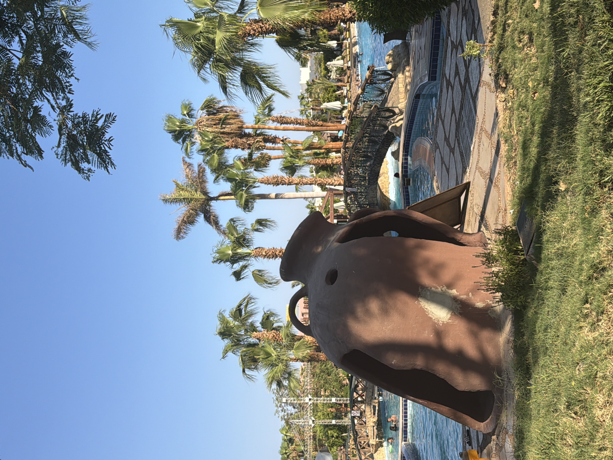 This is a daytime, eye-level shot of a resort pool area in Sharm El Sheikh, Egypt. In the foreground, a large, brown, sculptural slide is partially visible on the left side of the frame. It appears to be made of a molded material and has openings and a smooth, curved surface. To the right of the slide, a paved area with irregularly shaped stone tiles surrounds the pool. A metal bridge with decorative railings spans over the pool in the mid-ground. The pool itself is filled with clear blue water, and several people are visible in it, appearing to be swimming or relaxing. Palm trees are abundant throughout the scene, with lush green fronds reaching towards the clear blue sky. In the background, buildings and some scaffolding or structural elements are visible, suggesting the presence of resort infrastructure. The overall environment is sunny and appears to be a leisure setting.