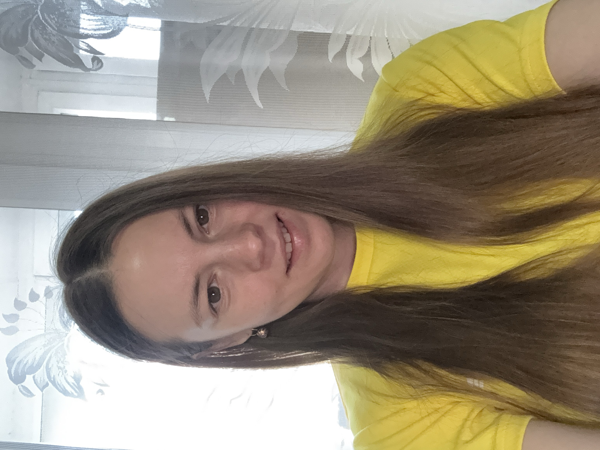 The image, rotated 90 degrees clockwise, captures a young woman taking a selfie indoors during the daytime. She has long, straight, dark brown hair, parted down the middle, that falls over her shoulders and a bright yellow short-sleeved t-shirt. Her head is tilted slightly, and she is looking directly at the camera with a subtle, closed-lip smile, showing a hint of her teeth. She has dark brown eyes and clear skin, and a small, round, metallic earring is visible on her left ear.
The background is dominated by a sheer, light-colored curtain, which appears white or light grey, adorned with a delicate floral or leaf pattern. Behind the curtain, a strong, bright light source indicates a window, suggesting it is daytime. Parts of a window frame are faintly visible through the translucent fabric. The woman appears calm and pleasant. No discernible text is visible in the image.