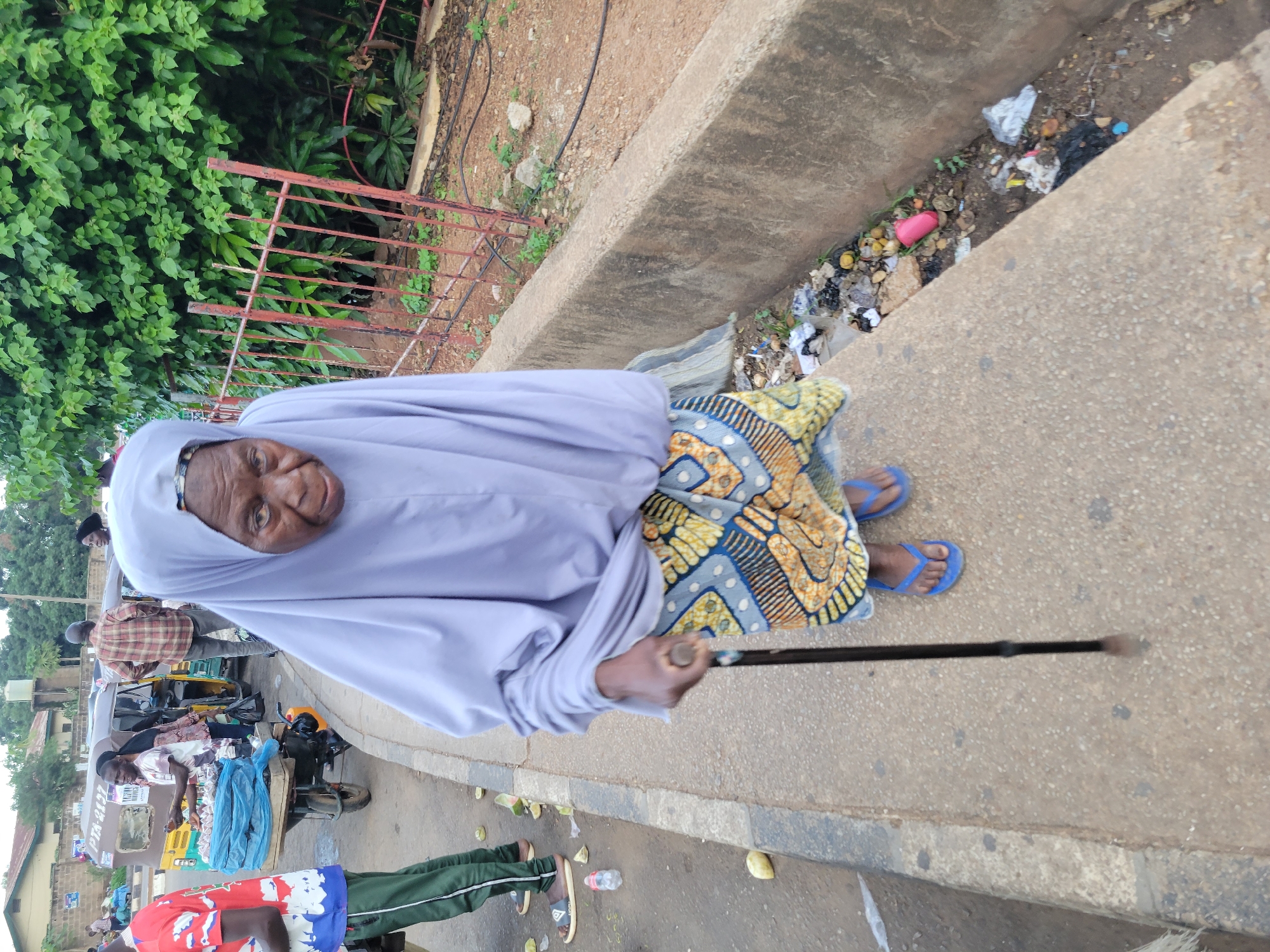 In the city of Barakin Lerawi, Nigeria, an elderly woman walks with a cane on a sidewalk next to a gutter filled with debris. She is wearing a light purple headscarf and a vibrantly patterned skirt. Her feet are adorned with blue flip-flops. To her left, on the street, a man in a red and white patterned shirt stands near a tricycle laden with goods, possibly a street vendor. Another individual in a red and white shirt and green pants is visible further down the street, near a blue plastic bag. The background features lush green foliage and a metal fence. The overall scene depicts a typical street environment in a Nigerian city, with the presence of street vendors, public transportation, and everyday pedestrian activity.