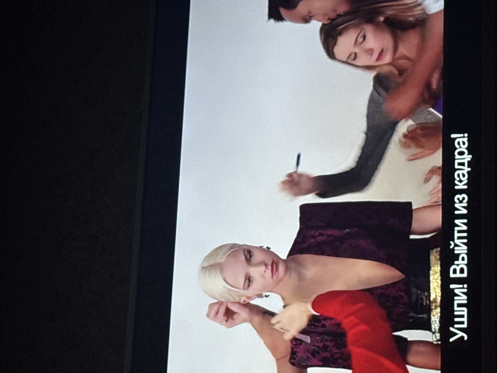 The image shows a scene from a video or movie being displayed on a screen. On the right side of the screen, there are two women being embraced by a man. In the foreground, a woman with blonde hair is visible. Russian text, "Ушли! Выйти из кадра!", is displayed on the right edge of the screen, translating to "Leave! Get out of the frame!". Location information about Mytishchi, Russia, is not evident in the image.