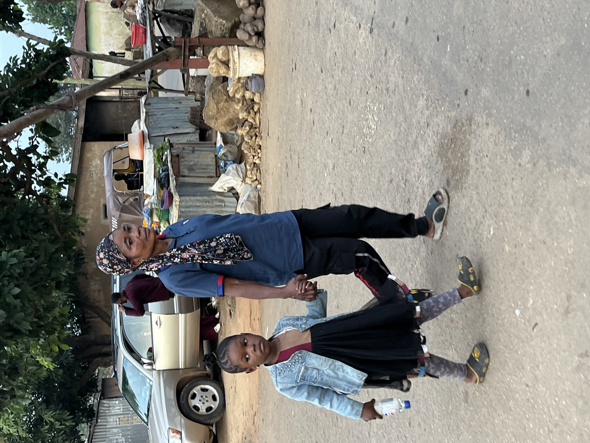 The image captures two individuals, an adult woman and a young girl, holding hands and standing on a paved road. The woman is wearing a blue shirt, a headscarf with a floral pattern, and dark pants. The girl is dressed in a denim jacket, a dark skirt, and leggings. They appear to be walking or posing for a photo together. In the background, there's a beige car with its door open, and a person is visible inside the car, leaning forward. Further in the background, there are structures made of corrugated metal, some sacks, and what appears to be a market stall with produce and other items. The scene suggests an outdoor setting, possibly a street or a marketplace, in Jos, Nigeria. The lighting indicates it is daytime. The overall mood seems neutral or perhaps a moment of connection between the two individuals. No visible text is discernible in the image.
