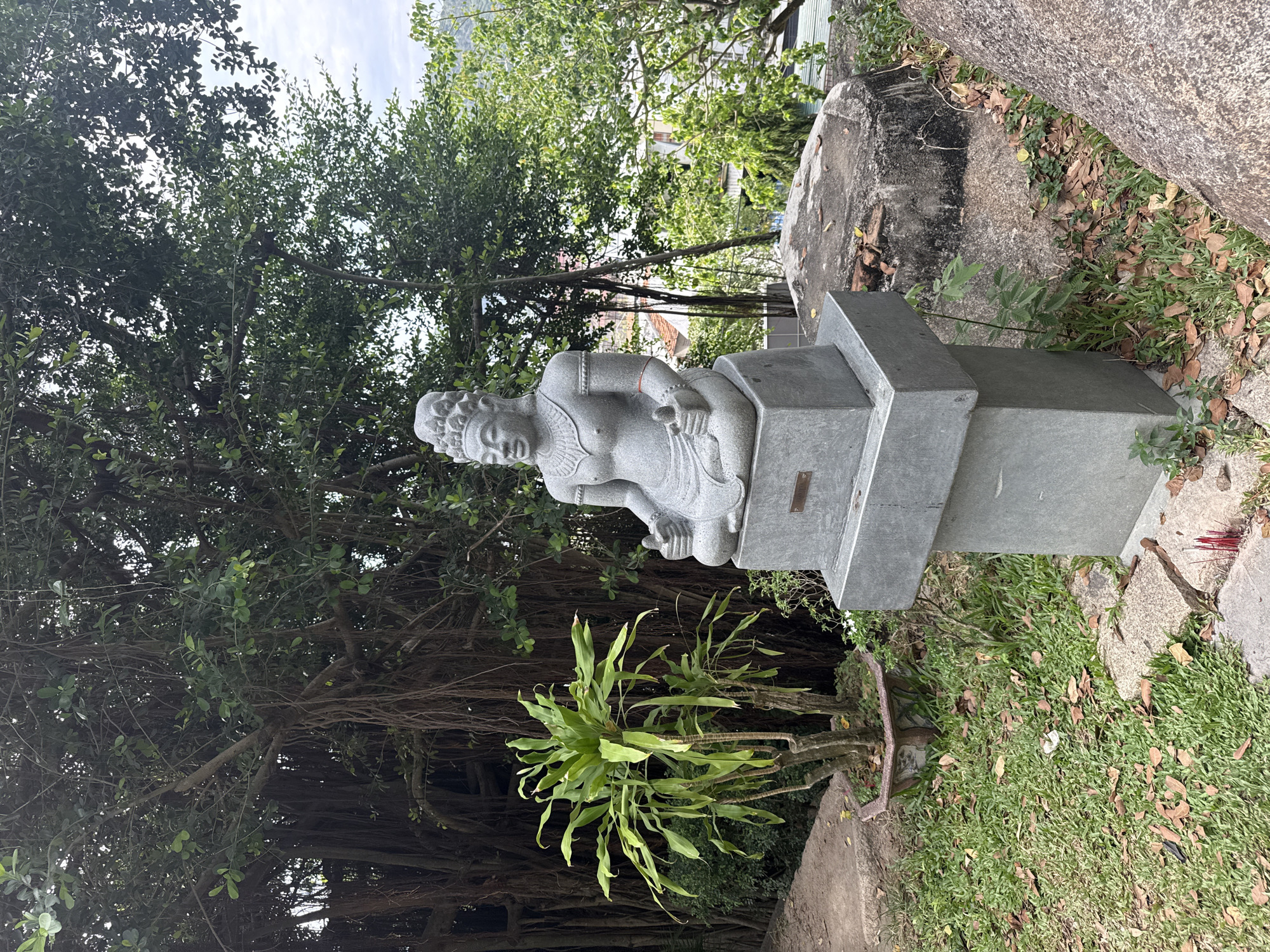 The image depicts a stone statue of a seated figure, possibly a deity or enlightened being, positioned on a stepped rectangular base. The statue is located in a garden setting with lush greenery. A large tree is behind and to the left of the statue, with aerial roots extending downwards. Smaller plants are visible at the base of the statue's pedestal. Incense sticks are nearby. The location is identified as Bac Nha Trang Ward, Vietnam. A large rock is to the right of the statue.