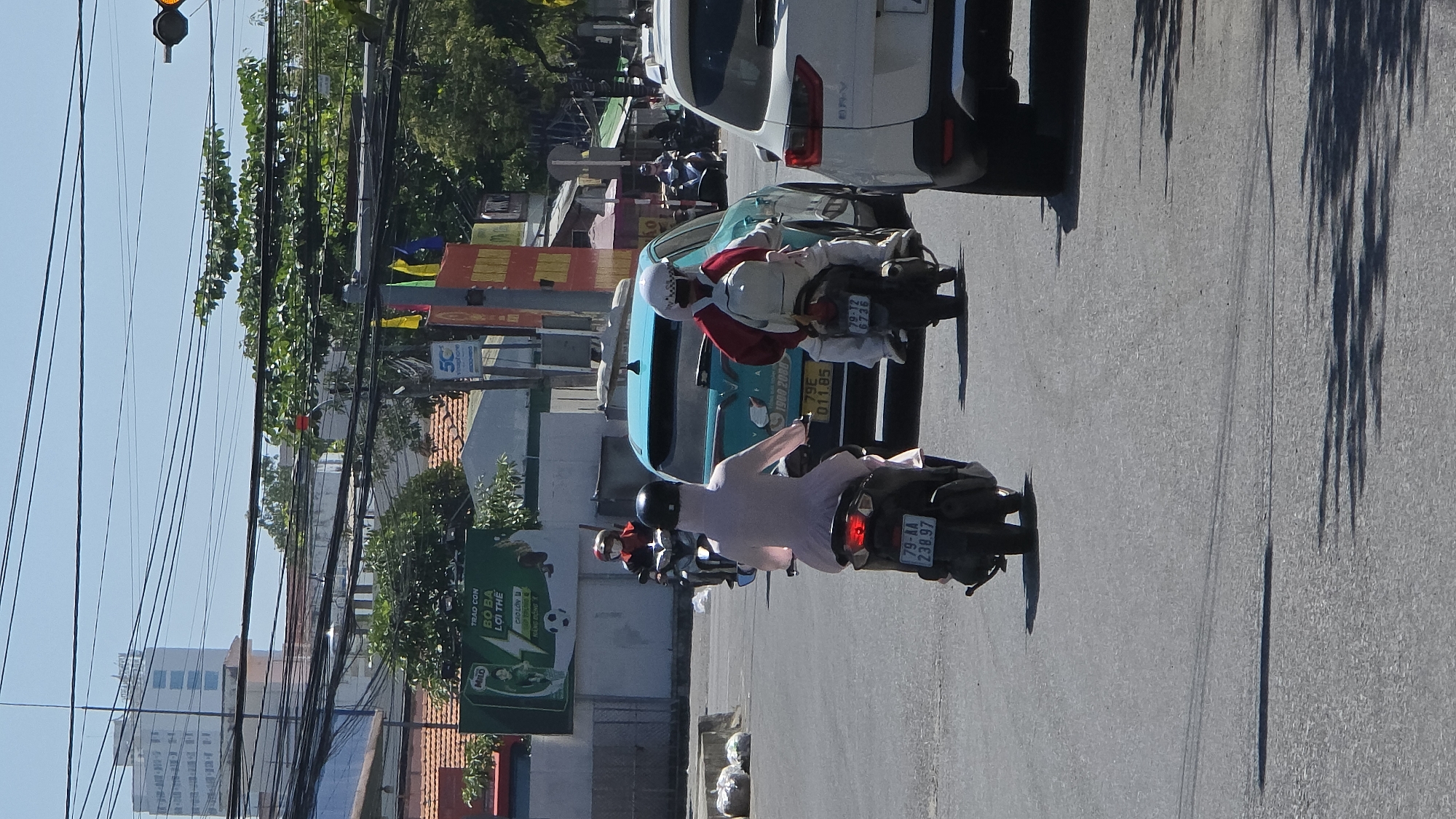 This image depicts an urban street scene under bright sunlight in Nha Trang, Khanh Hoa Province, Vietnam, identified by the "79" prefix on visible license plates. The asphalt road shows tire marks and tree shadows.
In the foreground, two motorbikes are visible from behind, traveling away from the viewer. The motorbike on the left is a dark scooter with license plate 79-AA 238.97, operated by an individual wearing a full-body, light pink sun-protection suit and a dark helmet. The motorbike on the right is also a dark scooter, license plate 79-T2 6736. Its rider wears white pants, a red long-sleeved jacket, a white helmet with a dark checkered pattern, and a white backpack.
Directly in front of the right motorbike is a teal-colored car, likely a VinFast Fadil, displaying license plate 79E 011.85 and a "VINFAST 1900 2323" decal on its rear window. To the right of the teal car, a partial view of a white SUV, identified as a Honda CR-V by its visible badging, occupies the adjacent lane. Further back, another motorbike with a rider in red and black attire is visible.
The background features a mix of urban structures. On the left, a green billboard for "MILO" with Vietnamese text "TRÀO CON BÓ BA LỢI THẾ" is prominent, above tiled roofs and a concrete barrier. On the right, storefronts with various signs, including a "5G Viettel" banner, line the street. A network of numerous black utility lines crisscrosses the upper portion of the frame, partially obscuring a clear blue sky and a yellow traffic light signal at the top right. In the far distance, a tall, white multi-story building with blue-tinted windows rises above the cityscape. Green foliage is scattered throughout the background.