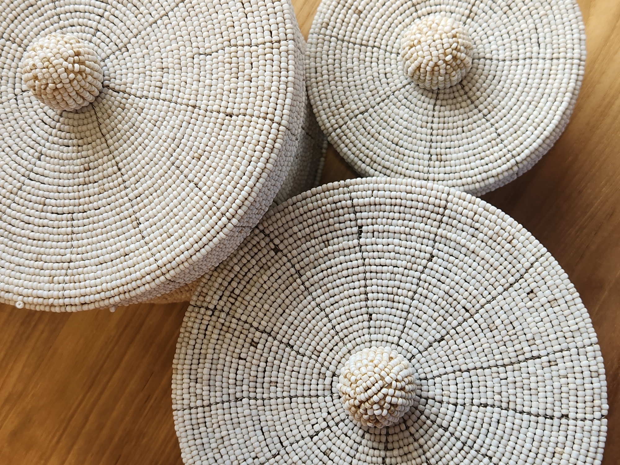 Three round, lidded containers, covered in small, off-white beads, are arranged on a wooden surface. Each lid has a small, spherical knob made of the same beads at its center. The containers are placed close together, with the lid of the largest container visible in the upper portion of the image. The other two containers are positioned below and to the left and right of the largest one, with their lids facing upwards. The wooden surface has a visible grain pattern. The scene is an indoor setting and does not provide any information about the city of Maitland, United States of America.