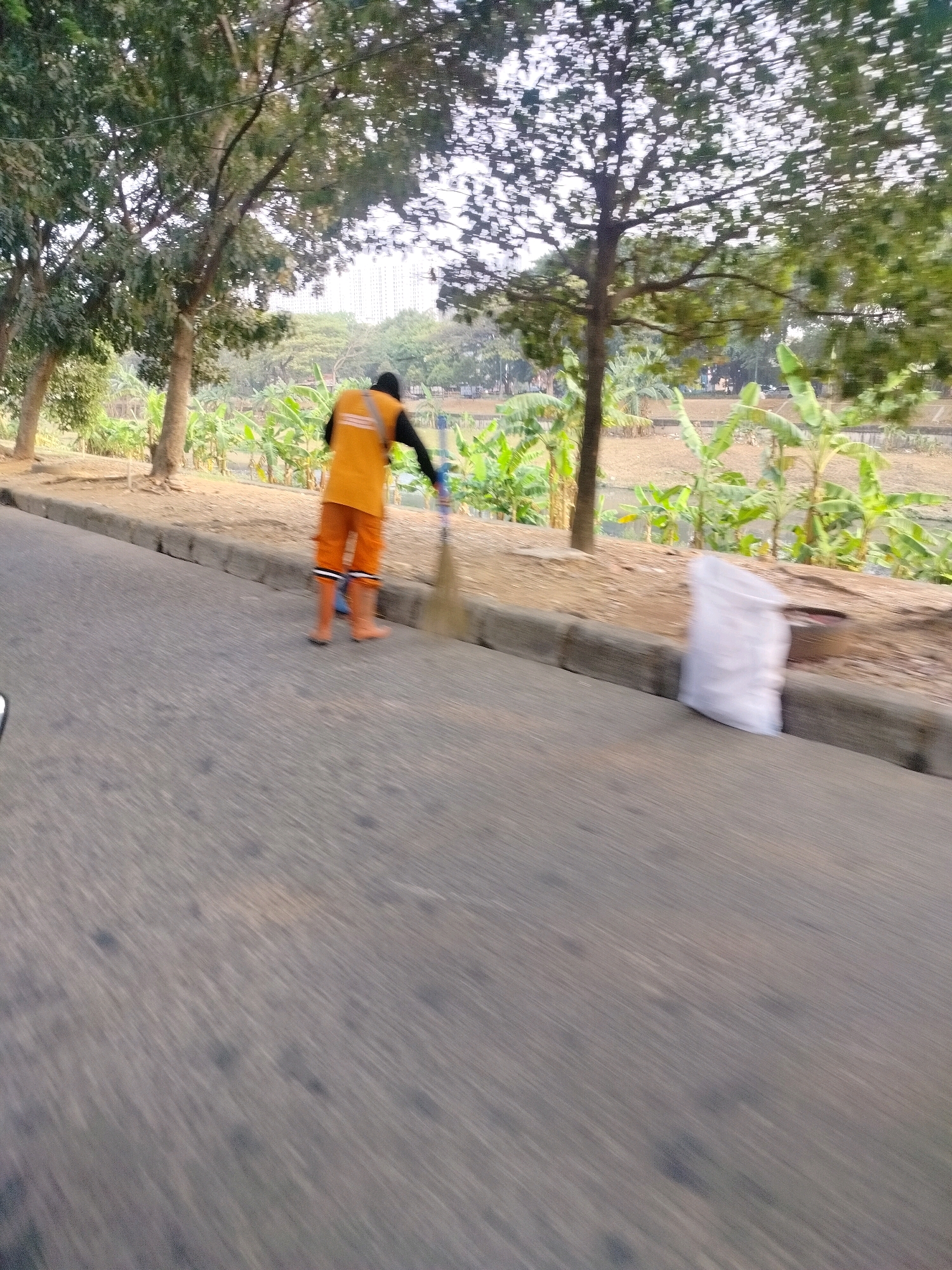A street sweeper wearing an orange vest and pants is sweeping the side of a road in East Jakarta, Indonesia. The street sweeper is holding a broom and appears to be collecting debris. A large white bag of trash is placed on the side of the road next to a dark-colored container. In the background, there are trees, plants, and a body of water. The image is blurry due to motion, suggesting it was taken from a moving vehicle. The overall scene depicts a typical urban environment with a focus on sanitation.