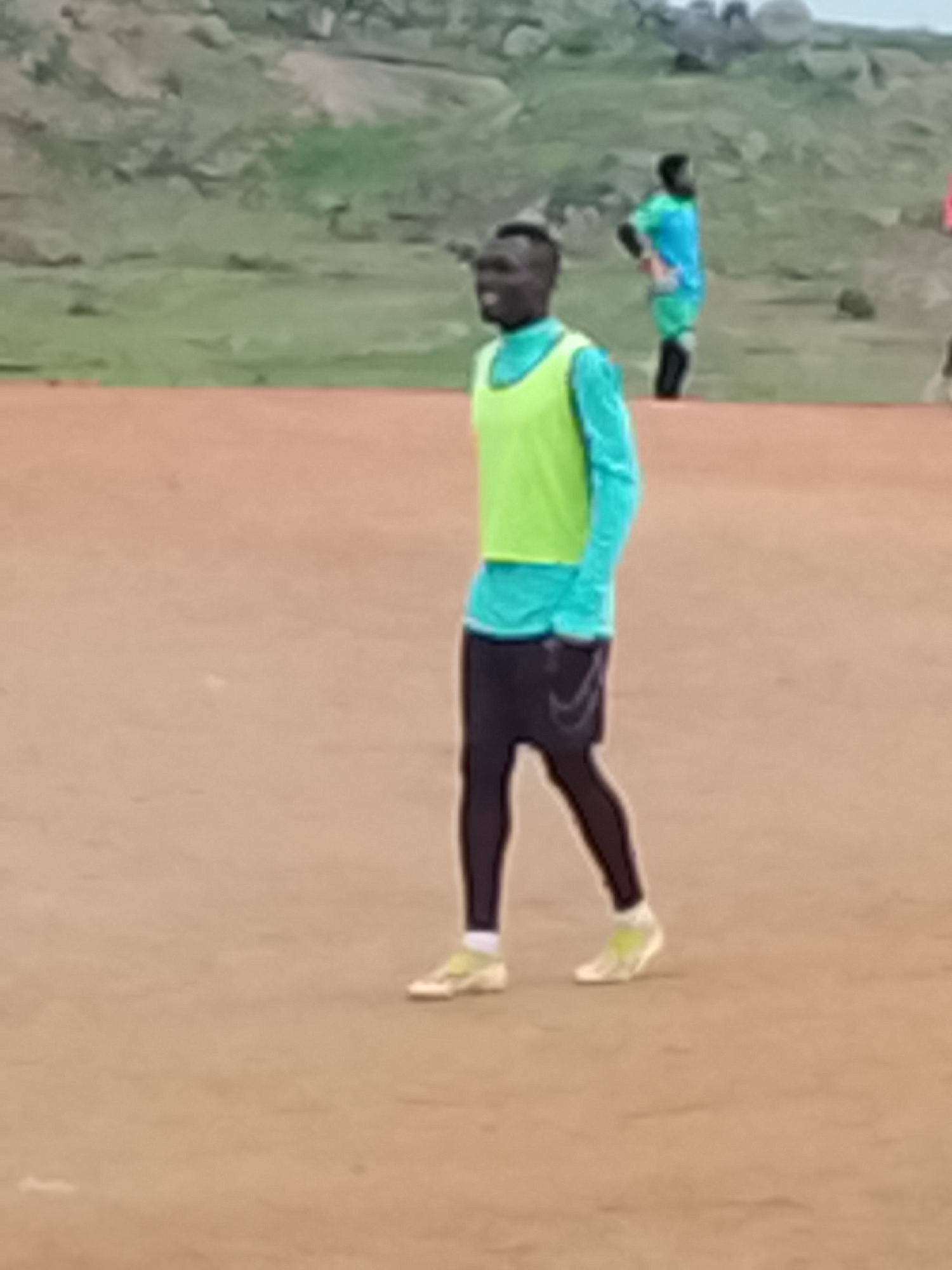 The image depicts a group of young men in athletic wear on a dirt field. In the foreground, a young Black man wearing a light blue long-sleeved shirt and a neon yellow vest stands facing slightly to the left. He has a shaved head and appears to be looking towards the left. He is wearing dark athletic shorts over black leggings and light-colored athletic shoes. In the background, another young Black man wearing a bright blue shirt and green shorts, with black socks, stands with his hands on his hips, looking to the right. A third person, partially visible, is wearing a pink shirt. The background shows a rocky, hilly landscape with sparse vegetation, suggesting an outdoor location. The overall impression is of a sports training session or casual game taking place in an open area. This scene could be indicative of a sports facility or a recreational space in a city like Jos, Nigeria, where such informal gatherings are common.