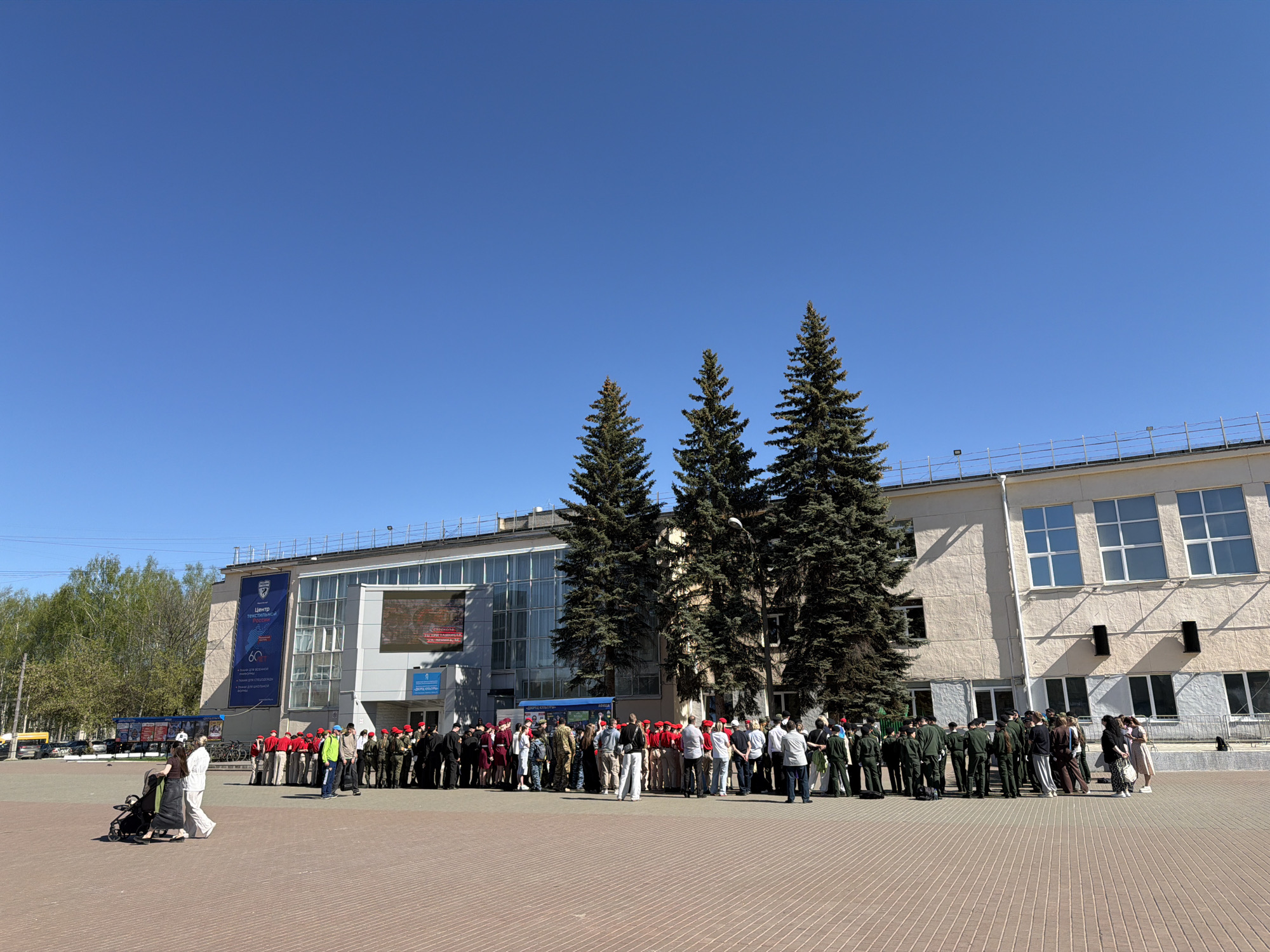 The image shows a group of people gathered on a brick-paved plaza in front of a building. The building is light colored with a mix of traditional and modern architectural elements, including large windows and signage. Three tall evergreen trees stand directly in front of the building. The people are dressed in a variety of clothing, with some wearing red hats and others in what appear to be military-style uniforms. There are also people in civilian clothes. A woman pushing a stroller walks across the plaza in the foreground. The sky is clear and blue. Based on the sign on the building that reads “Центр Творческих Технологий” (Center for Creative Technologies) and its 60-year anniversary banner, this appears to be the Center for Creative Technologies located on Lenin Street in the city of Chaikovsky, Russia.