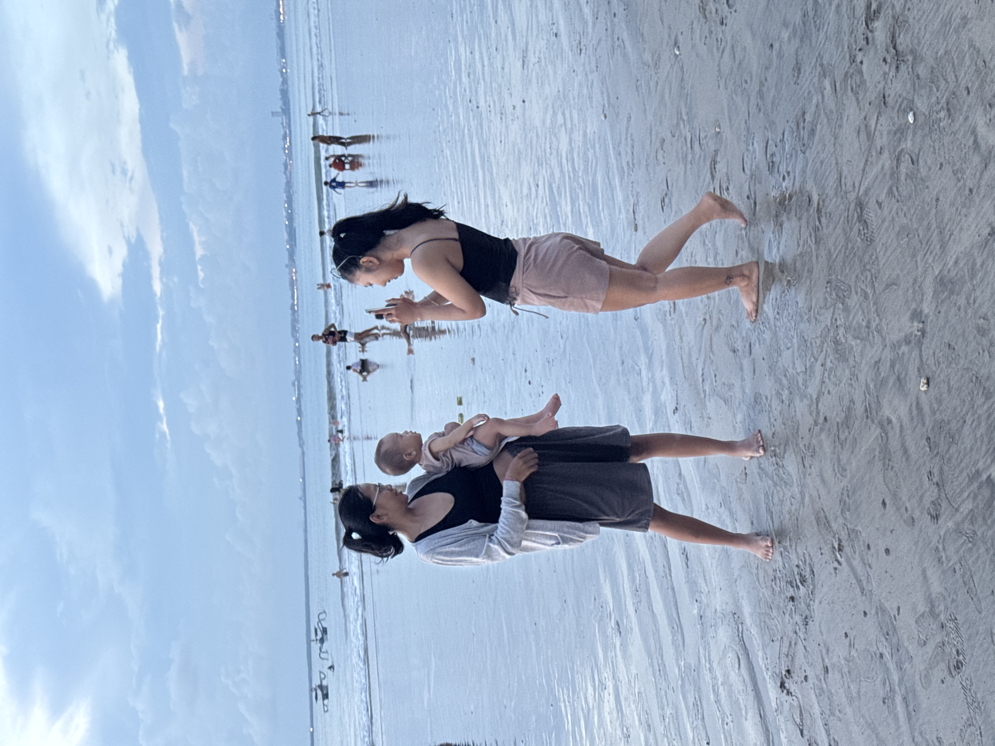 The image depicts a beach scene. One woman, wearing a black top and pink shorts, stands barefoot on the wet sand, looking down at her phone. Nearby, another woman holds a baby in her arms. She is wearing a black top with a grey skirt and a white cardigan, and is also barefoot. The baby is dressed in light pink clothing. In the background, there are other people walking along the shoreline and on the wet sand. The sky is visible above the water, with some clouds present. The location appears to be a beach, possibly in an urban area, but no city is definitively identifiable.