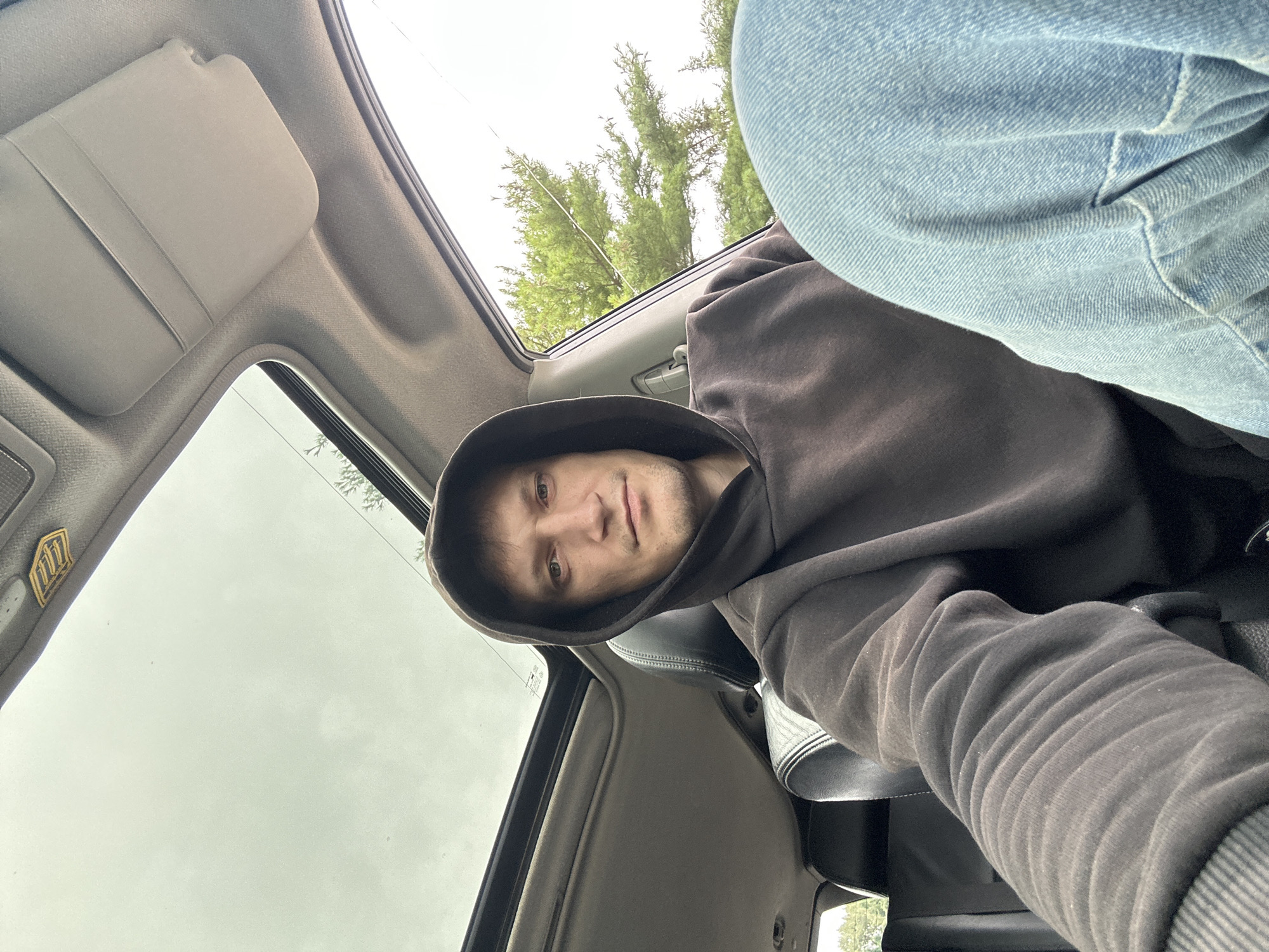 A man with short brown hair and light-colored eyes wears a dark brown hooded sweatshirt and light blue jeans. He is seated in a car, with his left arm resting on the center console. The car has a sunroof, which is open, revealing a cloudy sky. Trees and foliage are visible outside the car window to the right. There is a golden emblem with the number "777" on the ceiling of the car, above the sunroof. The scene appears to be outdoors, with the car parked. The location is identified as Makiv, Ukraine.