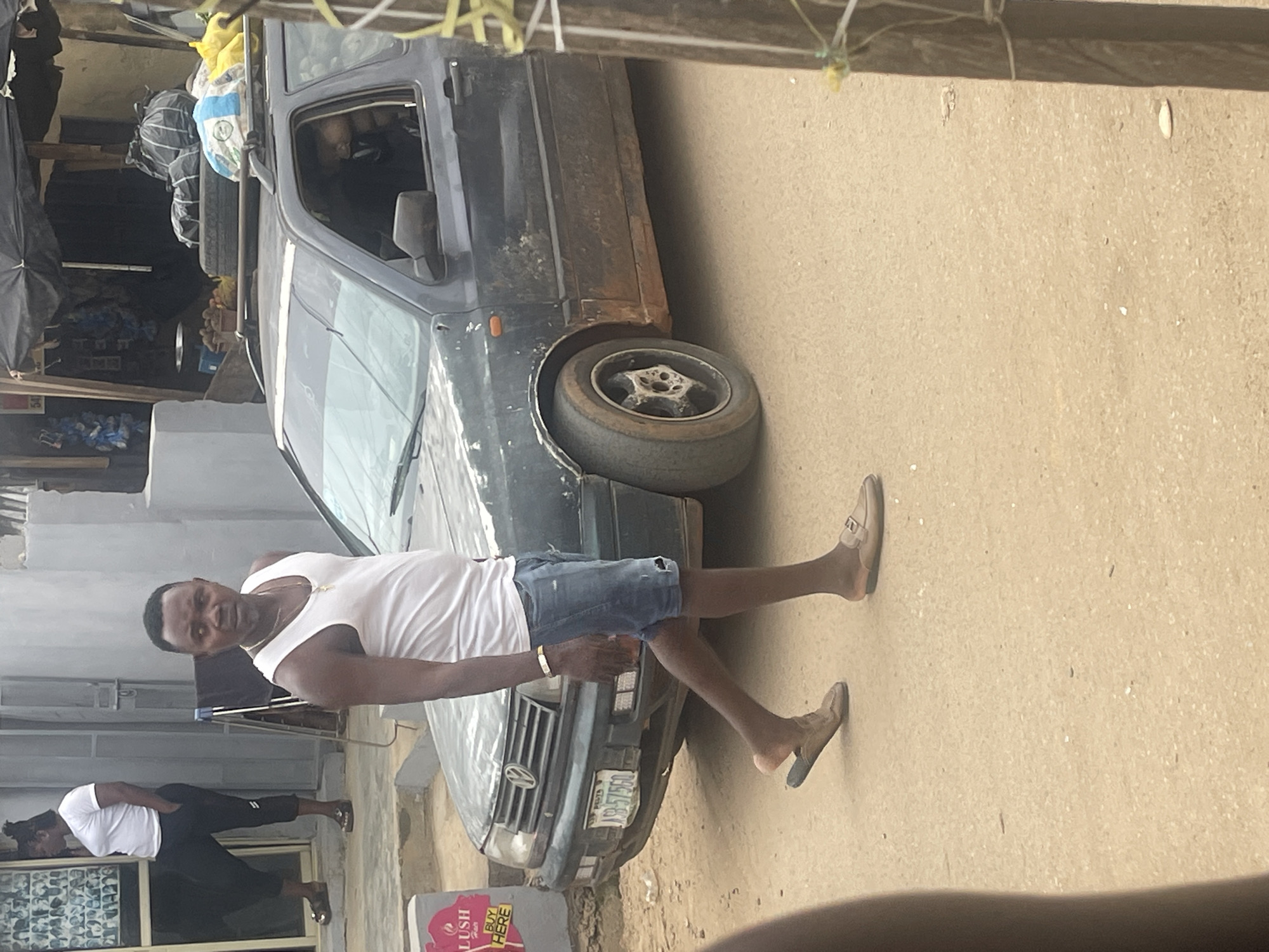 In Abuja, Nigeria, a man wearing a white tank top and denim shorts walks beside a dark, dirty station wagon. The man has short hair and is wearing a necklace. He is also wearing two different types of sandals. The car has luggage strapped to its roof and is parked on a dusty street. In the background, a woman in a white shirt and black pants stands near a building. The scene appears to be an outdoor market or roadside area.