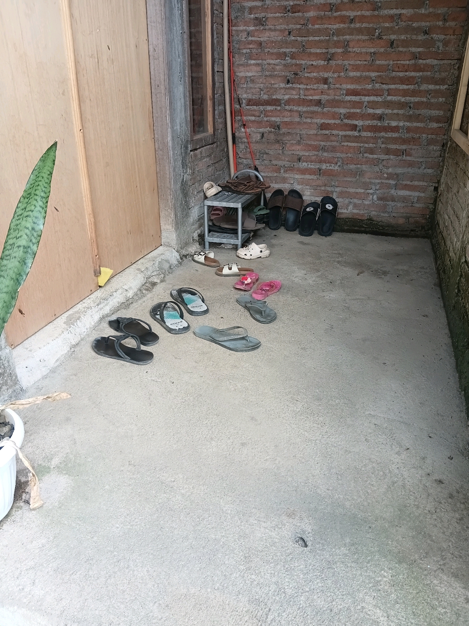 The image shows an outdoor area, likely a patio or entryway, in Cikal, Indonesia. The scene is dominated by a concrete floor and a brick wall. Various pairs of footwear, including flip-flops and sandals, are scattered across the ground and on a small metal shoe rack. A portion of a wooden door and a potted plant are visible on the left side of the image. The lighting suggests it is daytime.