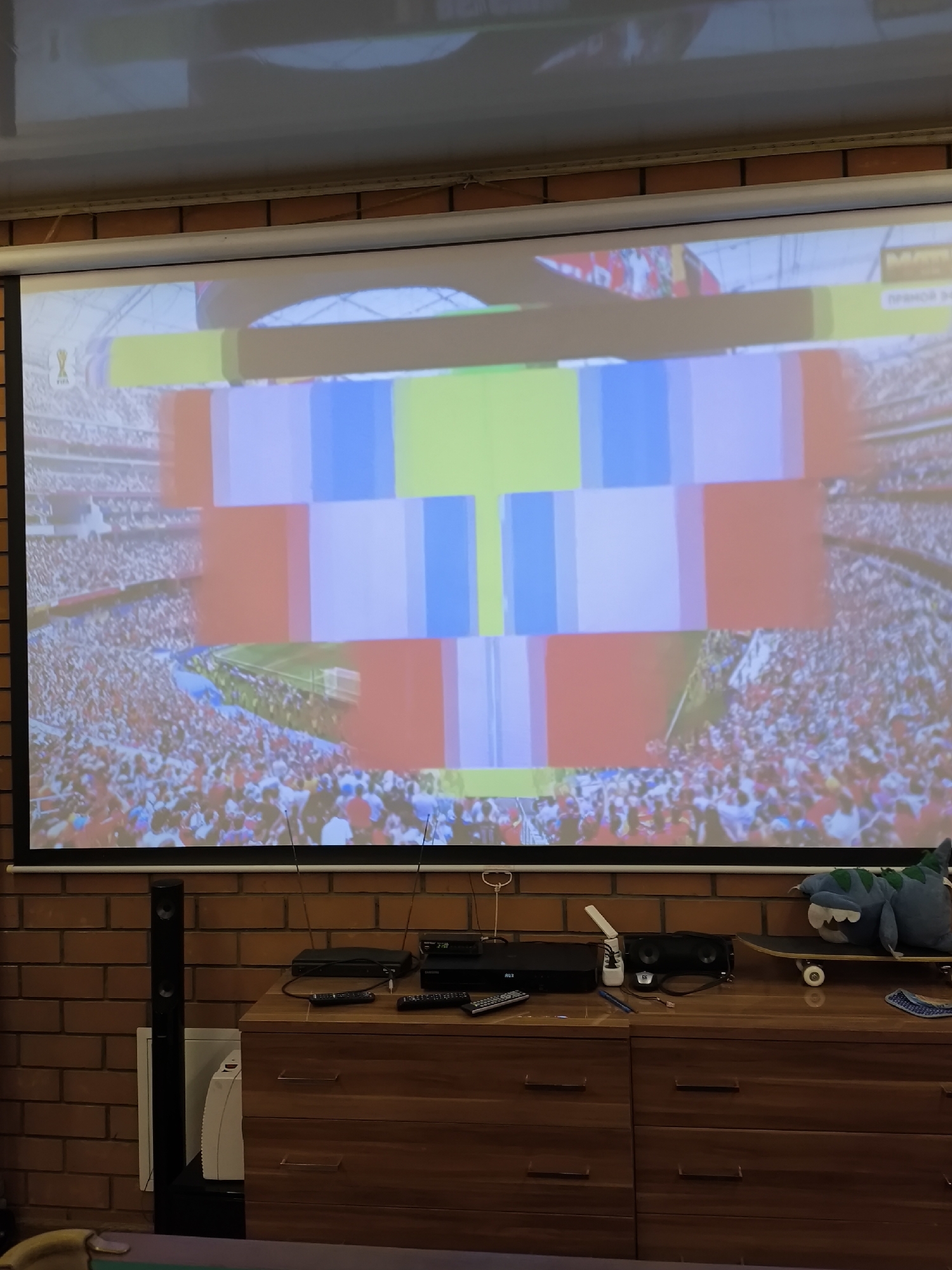 The image shows a projector screen displaying a distorted image of a stadium crowd and a colorful, pixelated overlay. Below the screen is a wooden dresser with various electronic devices and remotes. To the left of the dresser, a tall black speaker stands against a brick wall. The overall environment appears to be a living room or entertainment space. The location in the city of Pokrov, Russia is not discernible from the image.