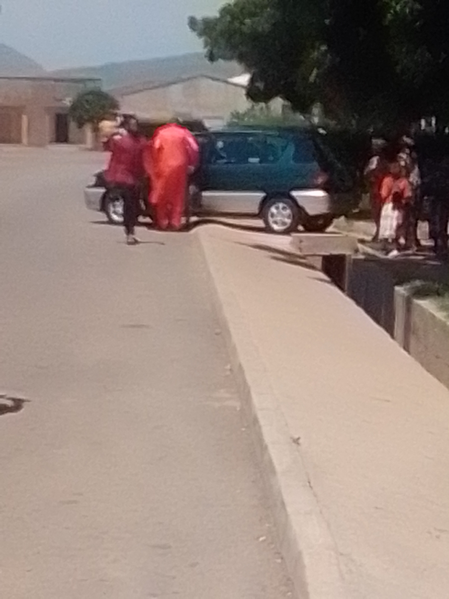 In Galadima, Nigeria, two individuals wearing bright red attire are interacting with the rear of a dark green minivan. One person, facing away from the camera, appears to be leaning into the vehicle, while the other, also in red and facing partially towards the camera, stands beside them. A third person, dressed in darker clothing and black pants, stands near the open rear door of the minivan. To the right of the minivan, several other individuals, including a child in a white garment, are visible, seemingly observing the situation. The vehicle is parked on a paved surface next to a raised concrete curb, with a dirt or gravel area beyond that. In the background, there are buildings and a cluster of trees, suggesting an outdoor, possibly semi-urban or residential setting. The sky is bright and clear.