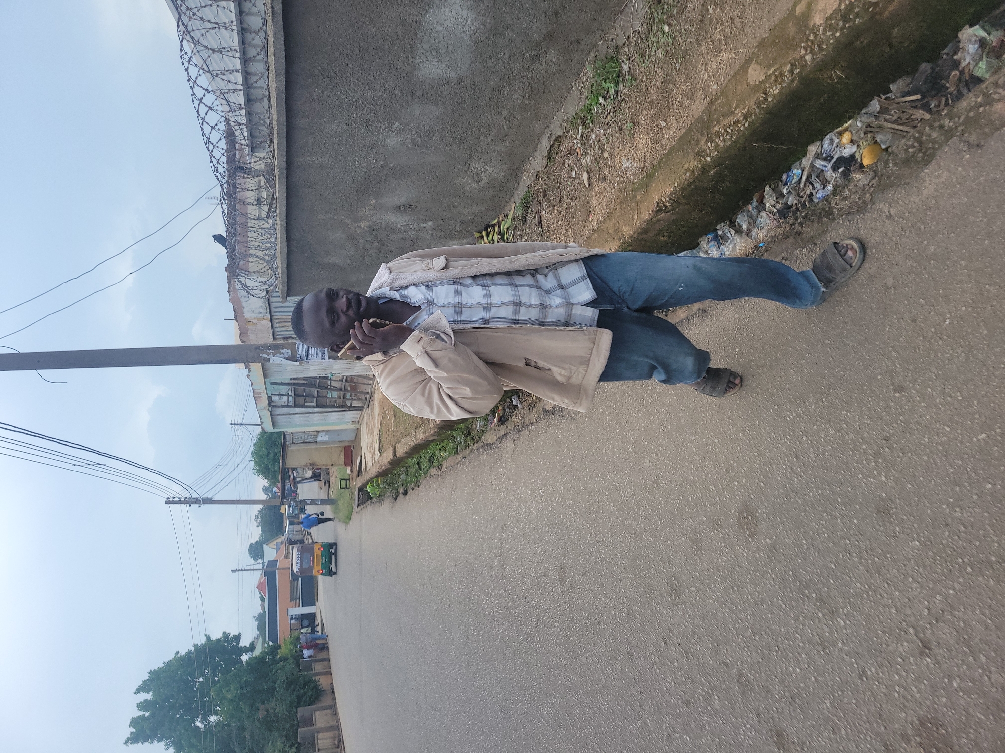 A medium shot captures a Black man in his 30s walking on an asphalt road in what appears to be a residential area in Jos, Nigeria. He is wearing a light-colored, collared jacket over a plaid shirt and blue jeans. His right hand is raised to his ear, holding a mobile phone, suggesting he is engaged in a conversation. He is wearing dark-colored sandals. The road is paved and appears to be a street with some vehicles and buildings in the background. To his right, a concrete drainage ditch is visible, filled with debris, including plastic bottles, wrappers, and other refuse. A stone wall topped with barbed wire fencing runs along the right side of the road, suggesting a boundary or security measure for an adjacent property. The sky is overcast, with visible power lines and poles dominating the upper portion of the frame. A few trees are present on the left side of the road, and the general environment appears to be a mix of residential and commercial structures.