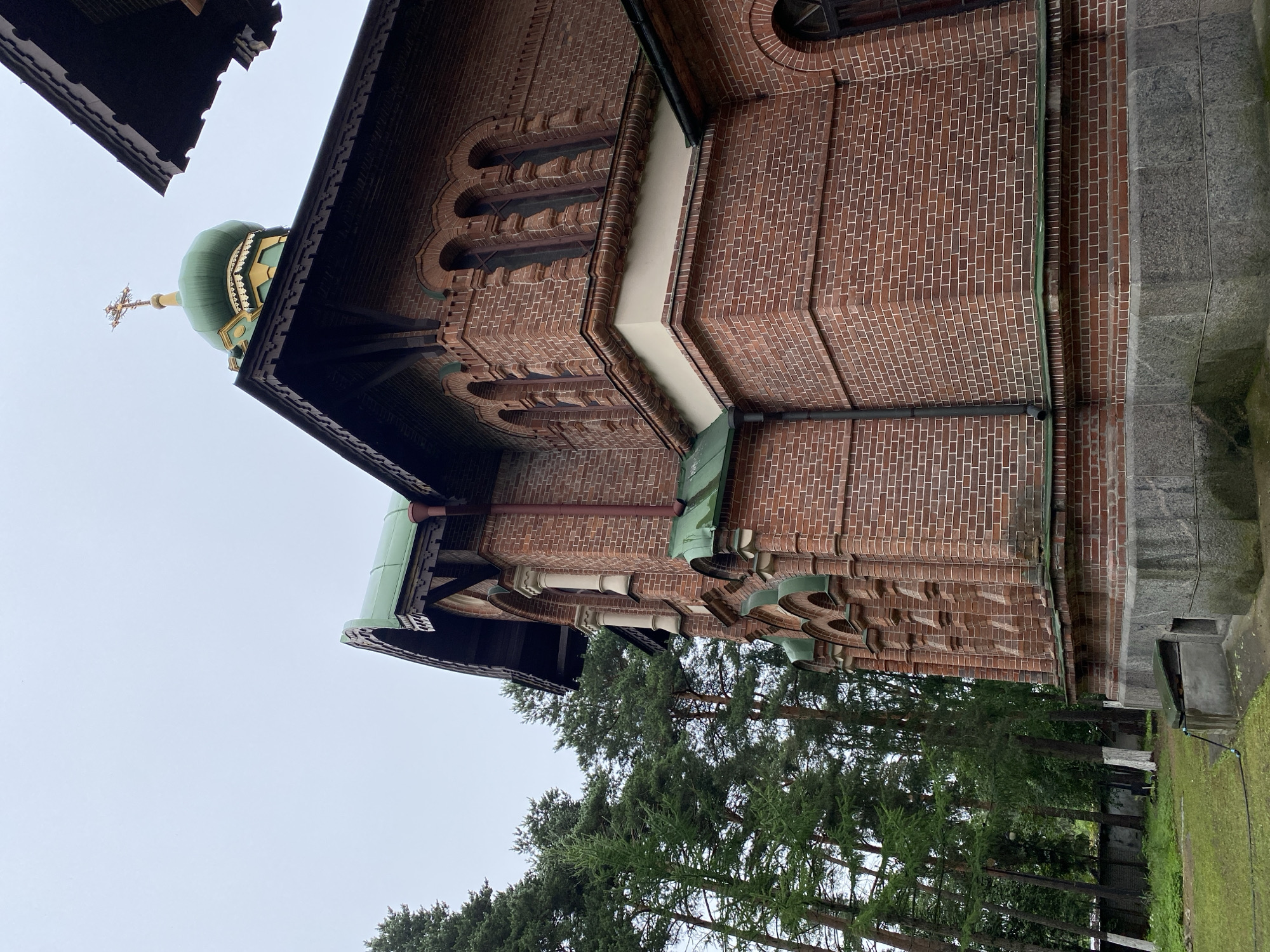 This is a medium-angle, outdoor, eye-level shot of the exterior of an Orthodox church in Priozersk, Russia. The church is constructed of red brick with dark brown, ornate wooden trim under the eaves and around the windows. A green dome with a golden cross on top is visible, along with sections of green metal roofing. The church has arched windows and decorative brickwork. Tall, green pine trees are to the left of the church, with a grey stone wall and some grass visible in the foreground. The sky is overcast and grey. No people are present.