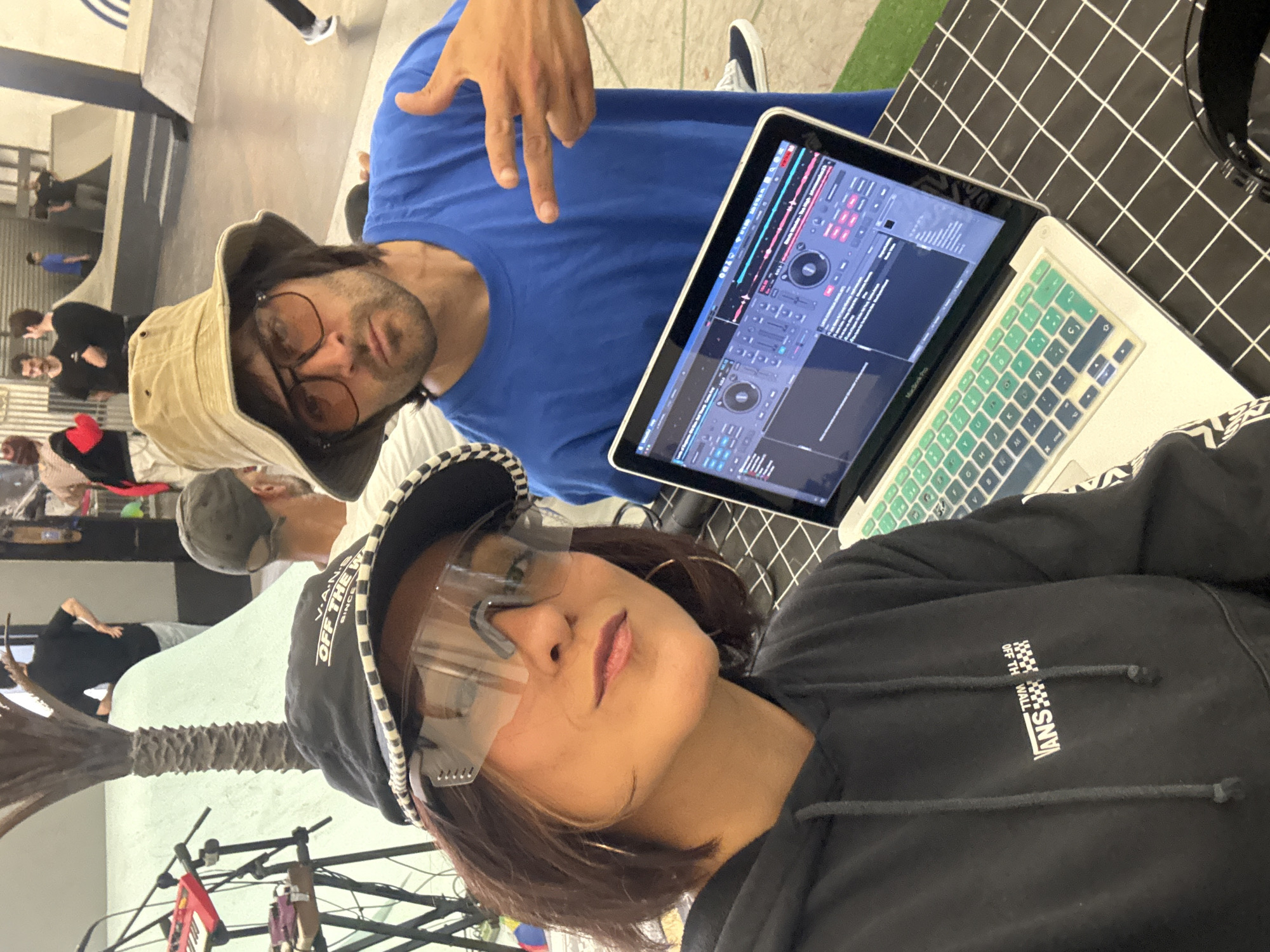 The image shows two people posing in front of a laptop. A man wearing a blue shirt, a bucket hat, and glasses is positioned slightly behind and to the side of a woman wearing a black hoodie, a cap with the "Vans" logo, and clear glasses. The laptop screen displays a DJ software interface. The background features a black and white checkered pattern, and a glimpse of a skate park environment is visible in the distance, including what appears to be a palm tree. The location is L'Hospitalet de Llobregat, Spain, suggested by contextual clues such as the setting resembling urban recreational spaces often found in cities.