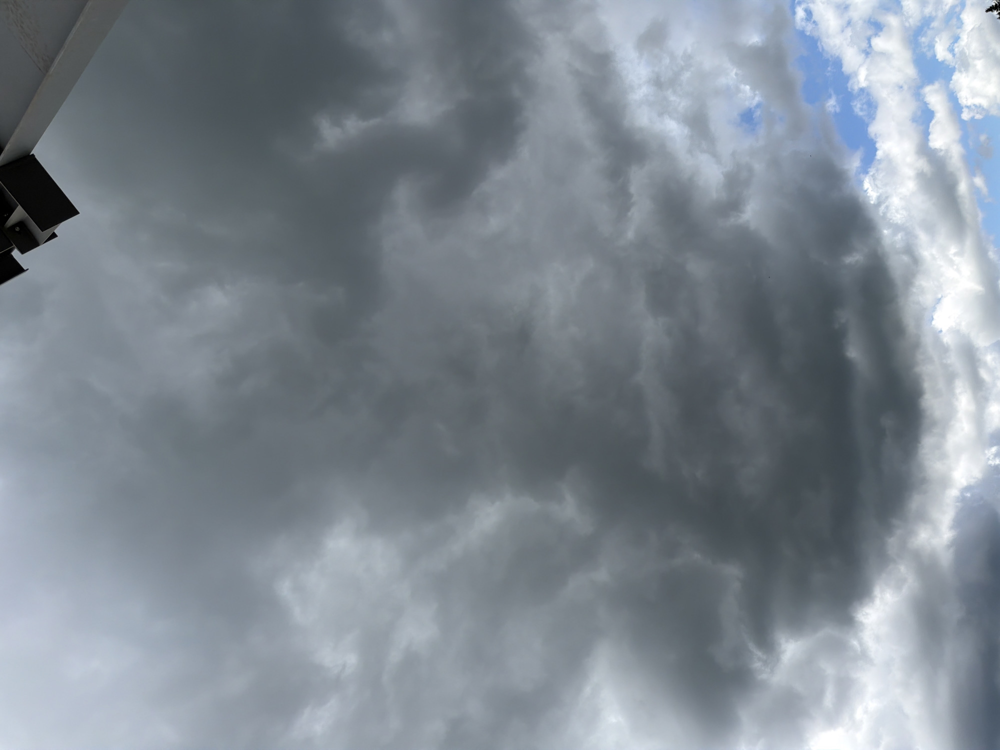 The image shows a dramatic sky filled with dark, ominous storm clouds. The clouds are dense and appear to be moving, with varying shades of gray and white. In the upper right corner of the image, a portion of a modern building's structure is visible, casting a shadow on the sky. The building appears to be made of metal and concrete. The lower part of the image reveals patches of clear blue sky with some lighter, fluffier clouds, indicating that the storm might be localized or passing. The scene suggests a turbulent weather event over the city of Janowice, Poland.
