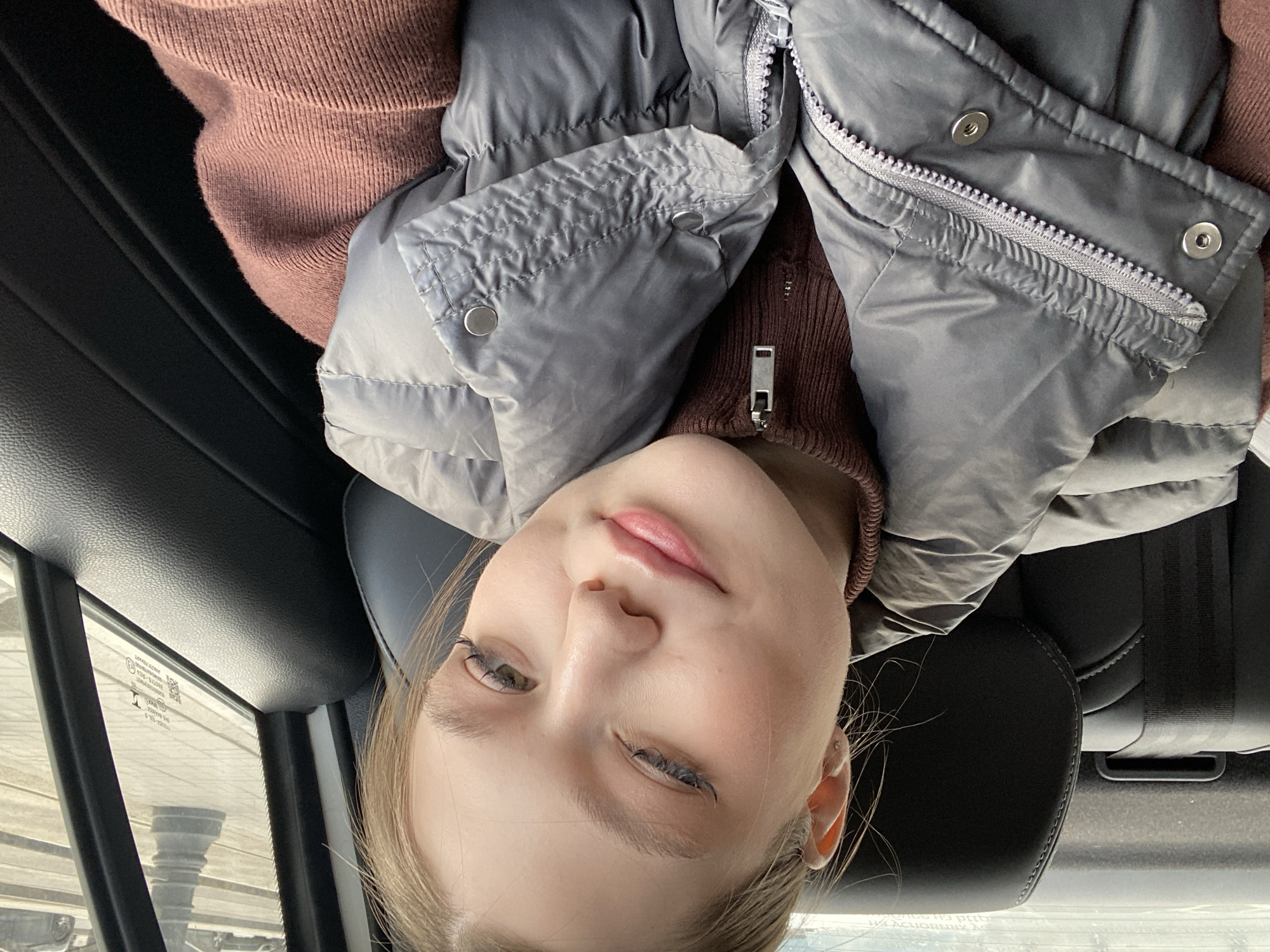 A fair-skinned young woman with blonde hair is in the backseat of a car. She is wearing a brown sweater and a gray vest. The woman is facing the camera with her eyes partially closed. A car window, with illegible inscriptions, is visible to her left, showing a street scene with a monument in the distance. The car seat and seatbelt are visible behind her. Based on the architectural style of the monument seen through the window, the location is most likely the city of Saint Petersburg, Russia.