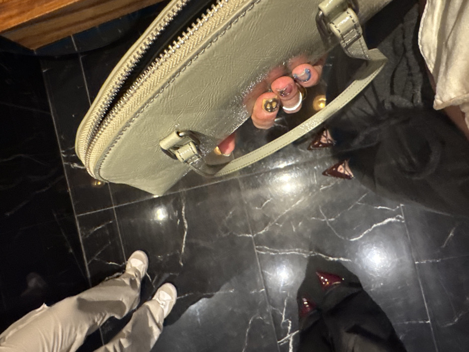 An overhead shot shows an open, light green handbag on a dark, marble-like floor. A pair of feet in light grey pants and sneakers are visible on the left. Inside the bag, toes with uniquely decorated nails and rings are visible. Scattered on the floor near the bag are several dark, triangular objects with white markings, along with dark shoes. Part of what appears to be white clothing is visible on the right.