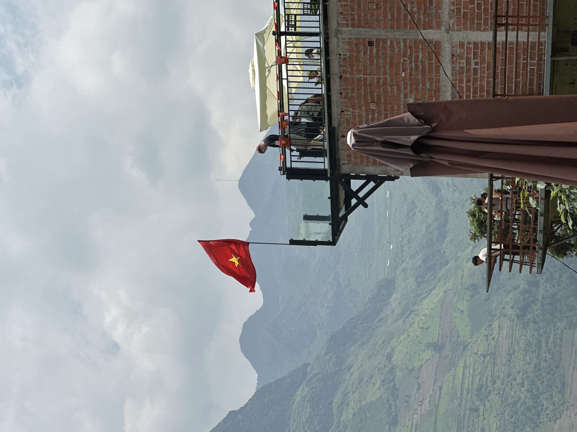 The image shows a scene from Mèo Vạc Commune, Vietnam. A Vietnamese flag is prominently displayed against a backdrop of mountainous terrain and a cloudy sky. A structure with a brick building and a glass-floored balcony extends from the mountainside. People are on the balcony, and a brown umbrella partially obscures the view.