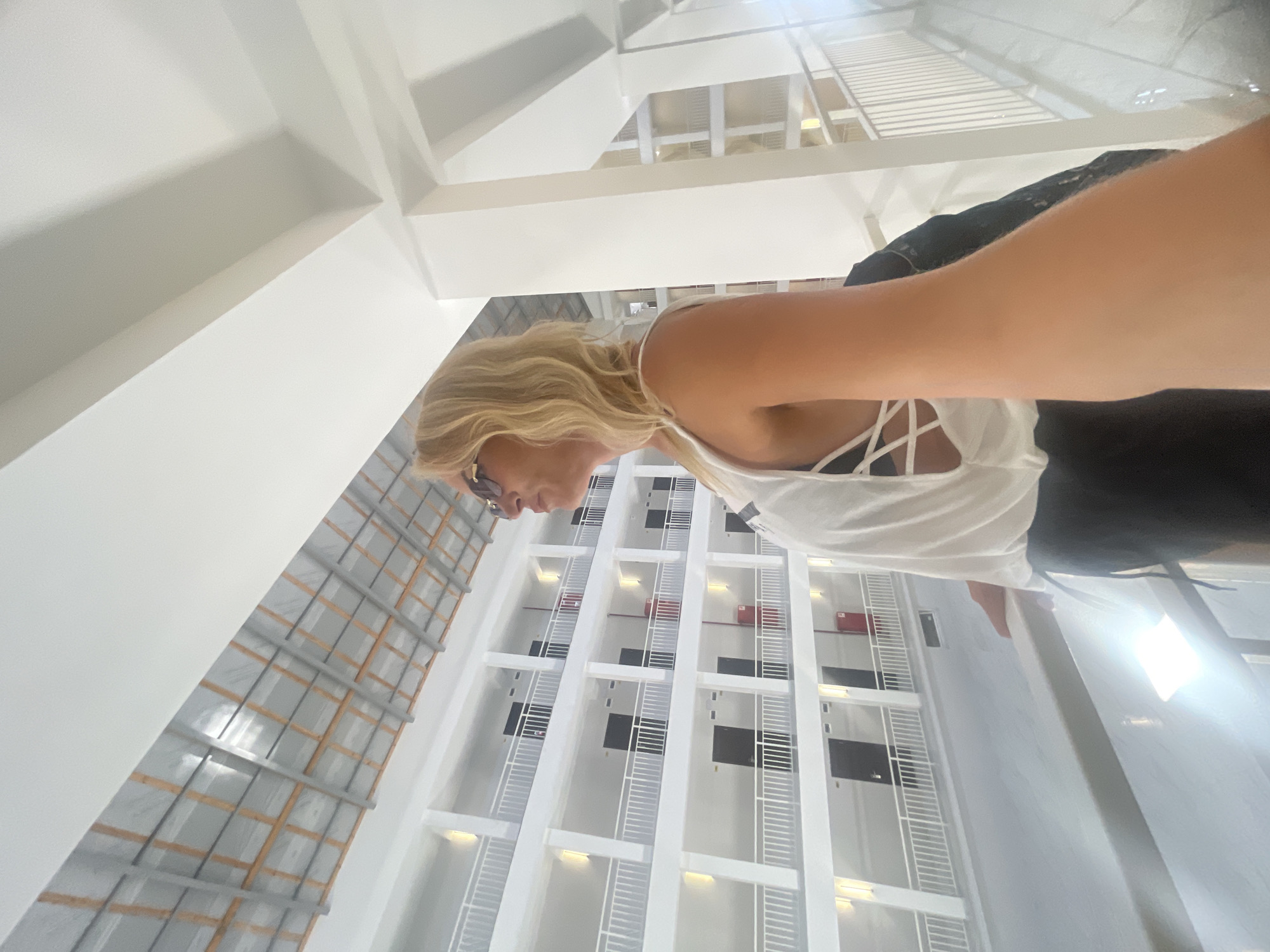 A woman with blonde hair and sunglasses, wearing a white sleeveless top and black pants, stands on a balcony overlooking a multi-story building with open corridors. The building has white railings and dark doors on each level. The woman is holding a black bag and extending her right arm, possibly taking a selfie. The environment appears to be an interior courtyard or atrium of a hotel or residential complex. The lighting is bright, with natural light entering from above and artificial lights illuminating the building's facade. The location is identified as Giachrypsh, Abkhazia.