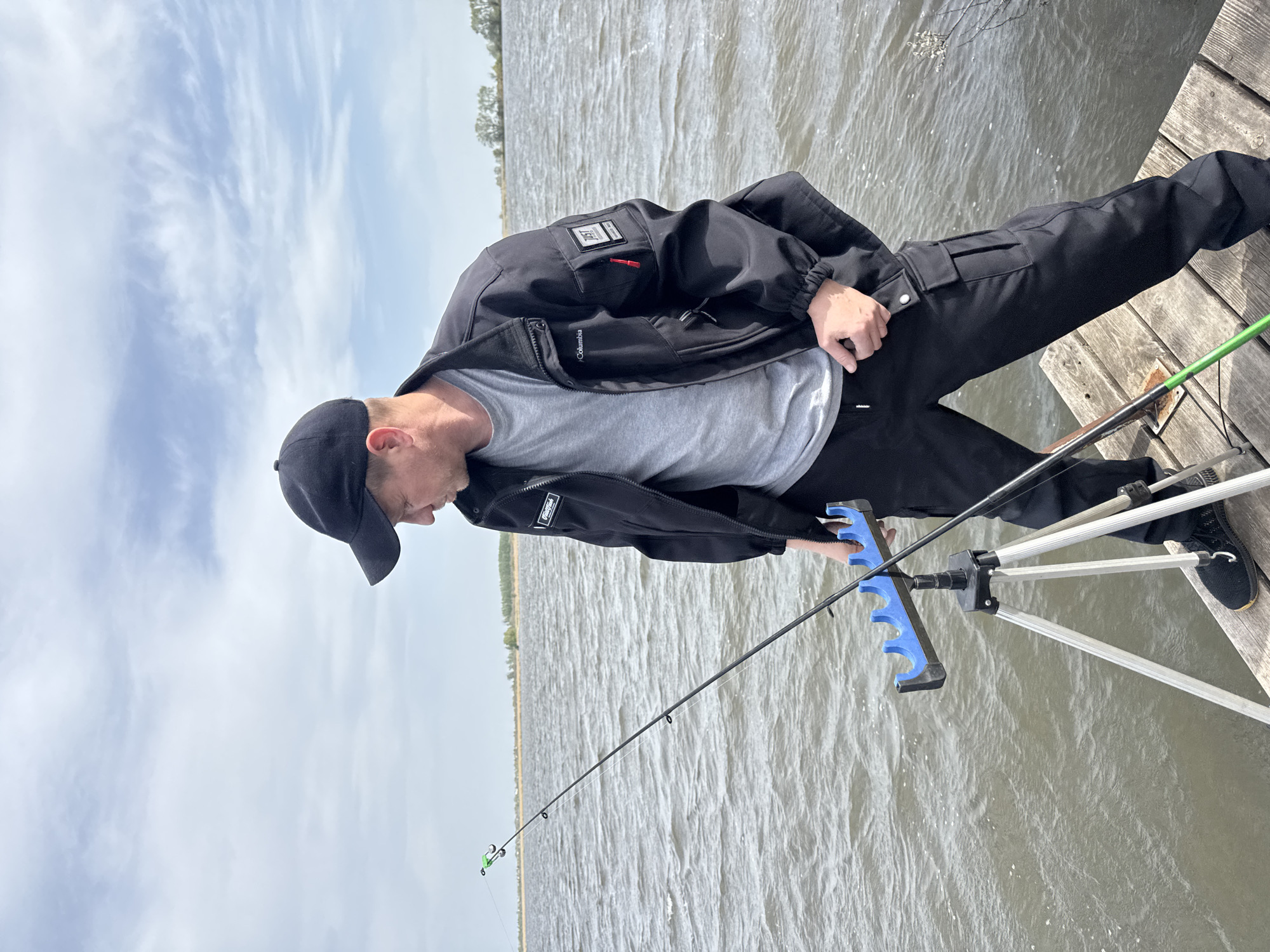 A man wearing a black jacket, gray t-shirt, black pants, and a black baseball cap is fishing on a wooden dock. He is holding a fishing rod with a blue rod rest attached to a silver tripod. The location appears to be a body of water with land visible in the distance under a partly cloudy sky. The location in the city is Unknown, Unknown.