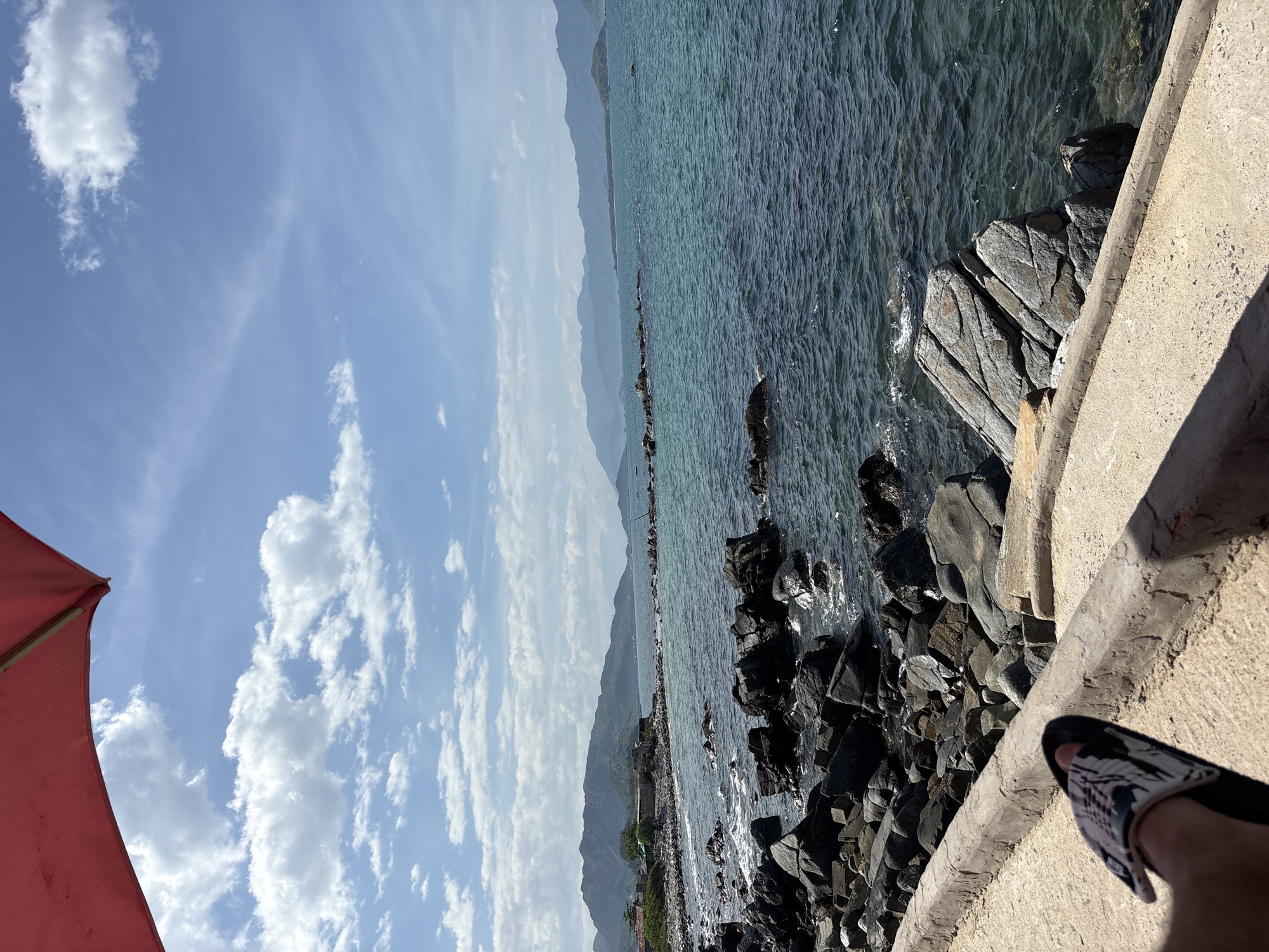 A person's foot wearing a sandal rests on a concrete ledge overlooking a rocky coastline in Vĩnh Lương, Vietnam. The sea is calm and blue, with gentle waves lapping against the dark rocks. In the distance, lush green mountains form a scenic backdrop against a sky filled with white, fluffy clouds. A portion of a red umbrella is visible in the upper left corner of the frame. The overall impression is one of a peaceful day at the beach.