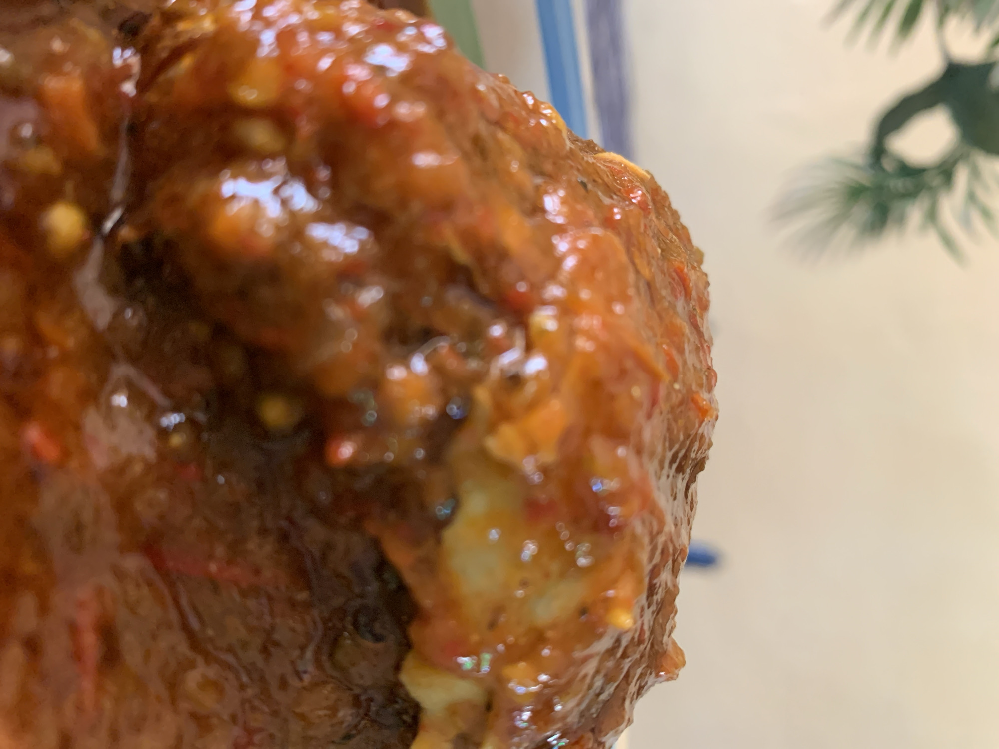 The image displays a close-up, shallow-depth-of-field photograph of a food item, located in New Stadium, Nigeria. The central subject is a solid, irregularly shaped item, likely a protein source, completely enveloped in a thick, viscous, reddish-orange sauce. The sauce exhibits a textured surface, with visible small red and orange particulate matter. A distinct layer of creamy yellow material, consistent with the albumen and yolk of a hard-boiled egg, is partially visible beneath the sauce on the upper-right portion of the food item. The base of the food item rests within a container, indicated by a blue and white striped pattern visible along the lower-left edge. The background consists of a uniformly blurred, light-colored surface, with a heavily out-of-focus dark green leafy plant visible in the upper-left corner. No human subjects, animals, or explicit actions are present within the frame. The scene is illuminated by soft, ambient light.