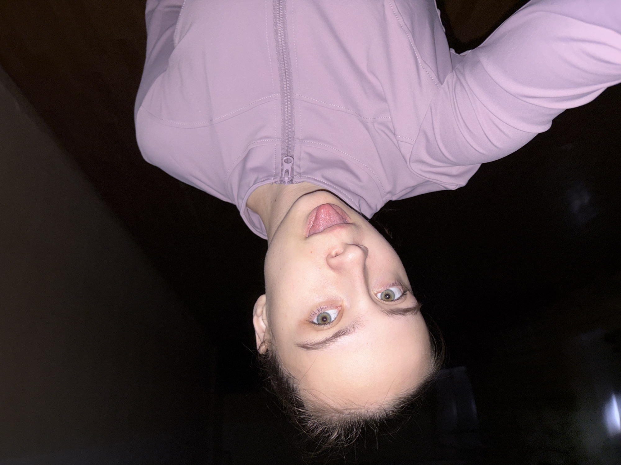 A young woman is making a silly face at the camera with her tongue sticking out. She is wearing a light pink zip-up jacket. The background is dark, suggesting low light conditions. The location is not discernible from the image. There is no explicit evidence or indication of the city of Chaikovsky, Russia in the image.
